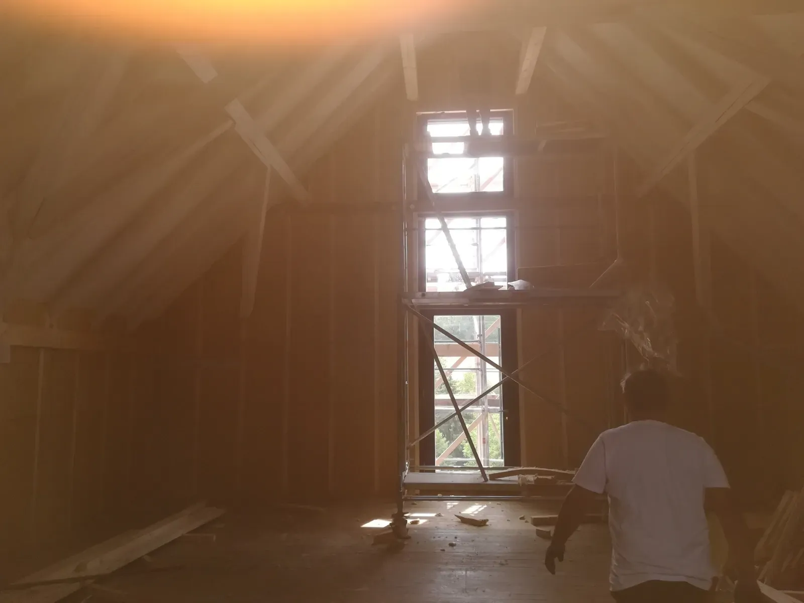 Staubiger Dachboden mit freiliegendem Fachwerk, Sonnenlicht fällt durch ein Fenster, und eine Person in einem weißen Hemd hockt nahe dem Boden.