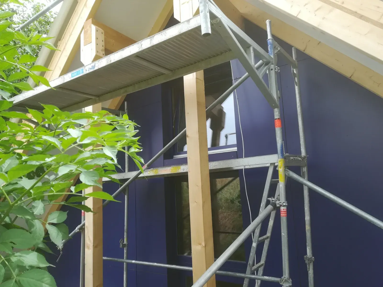 Ein Baugerüst neben einem im Bau befindlichen Haus, mit freiliegendem Holzfachwerk und blauen Wandpaneelen.