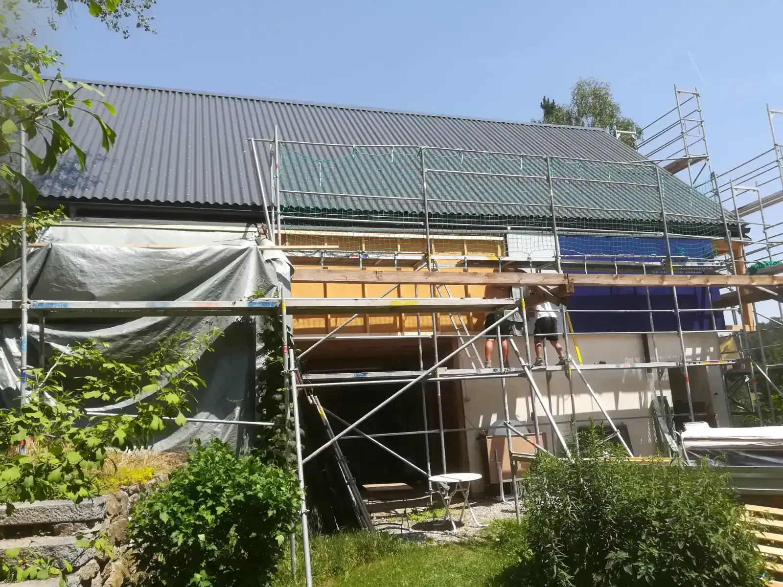 Haus im Umbau mit Gerüst und grauem Dach, umgeben von Grün