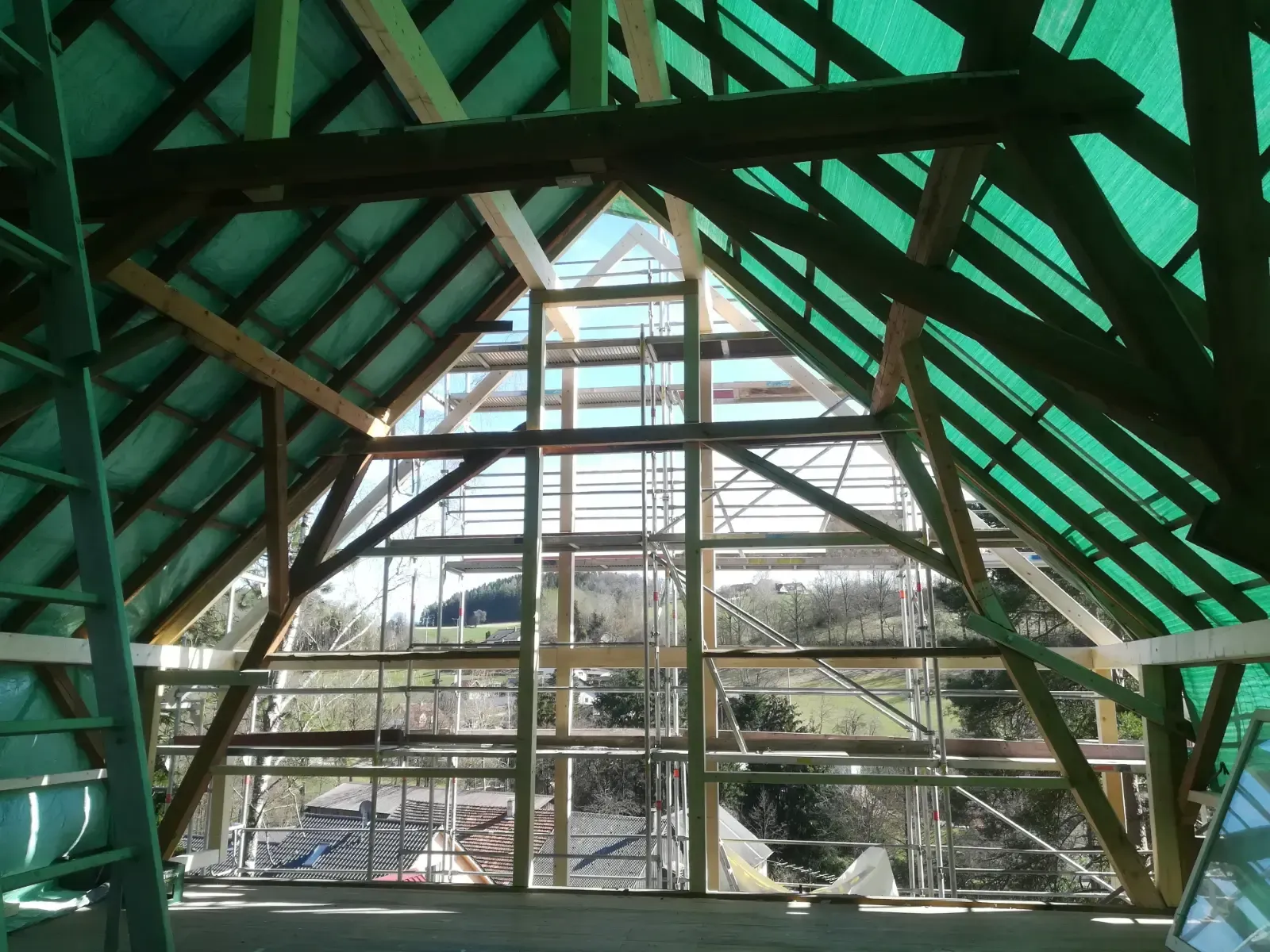 Innenansicht einer im Bau befindlichen, mit Grünrahmen versehenen Holzkonstruktion mit Dach, durch deren Fenster Gerüst und Hanglage sichtbar sind.