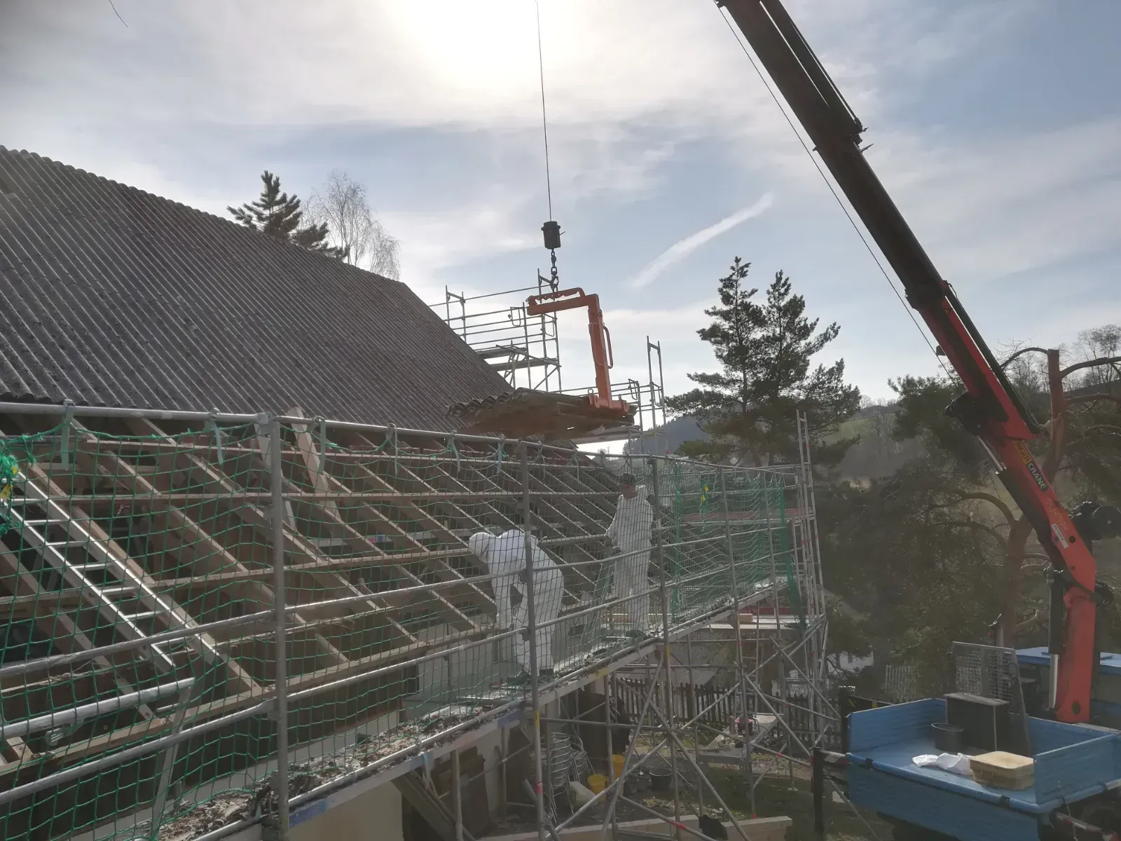 Ein Kran hebt den Dachstuhl über ein im Bau befindliches Haus mit Gerüst und Netz an.