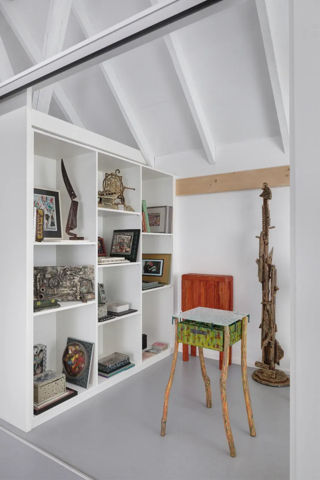 Helles, weißes Loftzimmer mit Einbauregalen, farbenfroher Dekoration, einem kleinen Hocker und einer geschnitzten Holzskulptur