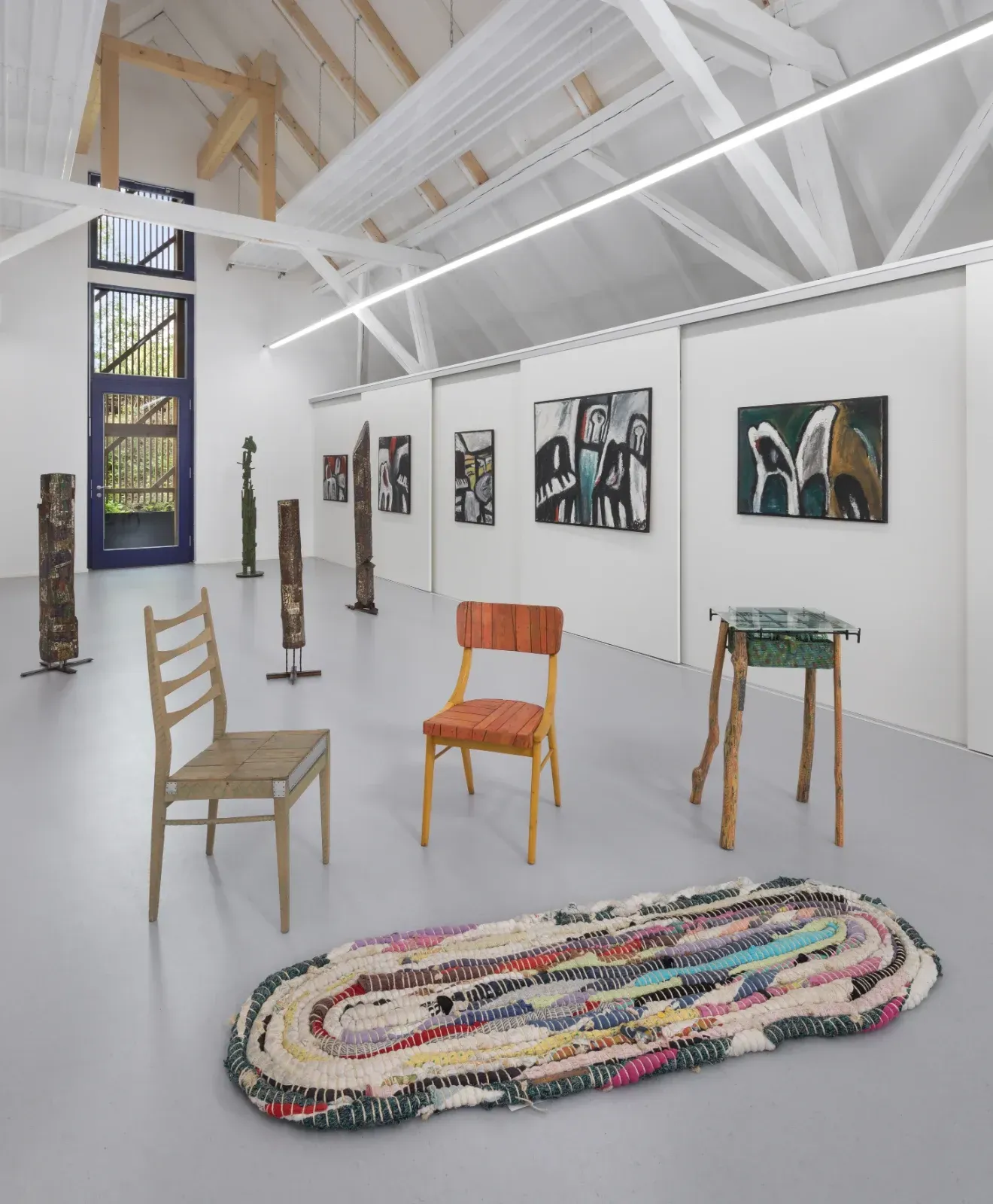 Kunstgalerie mit weißen Wänden, farbenfrohen Stühlen, gerahmten Gemälden und einem gewebten ovalen Teppich unter freiliegenden Balken.
