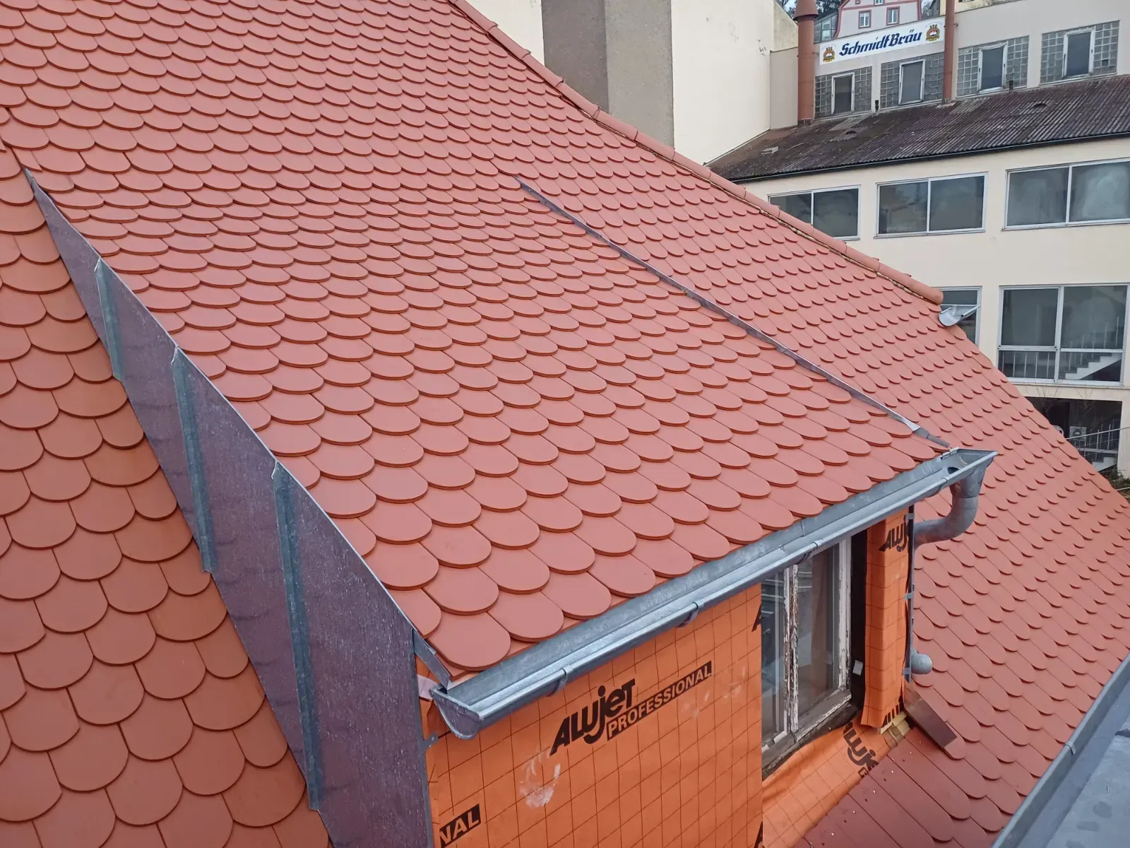 Ein mit roten Ziegeln gedecktes Schrägdach mit Dachgaube und orangefarbener Wand neben einem Gebäudeinnenhof
