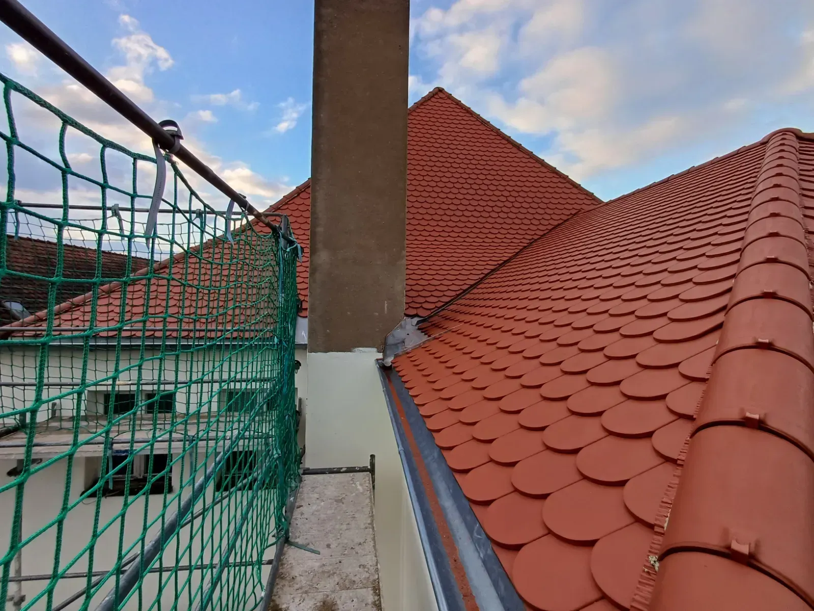 Rote Ziegeldächer und ein Schornstein neben grünen Sicherheitsnetzen an einem im Bau befindlichen Gebäude.