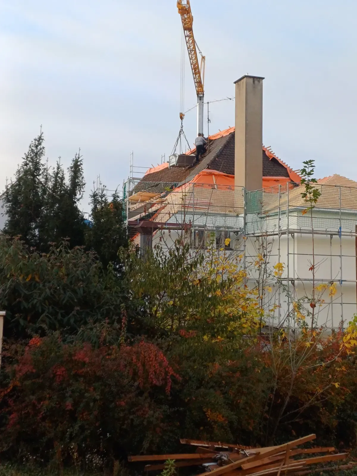 Hausdach wird repariert; im Vordergrund sind ein orangefarbener Kran, ein Schornstein und Bäume zu sehen.