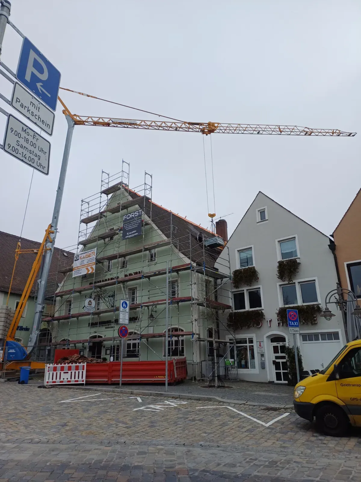 Ein eingerüstetes Gebäude im Bau auf einer Kopfsteinpflasterstraße, darüber ein Kran und in der Nähe geparkte Fahrzeuge.