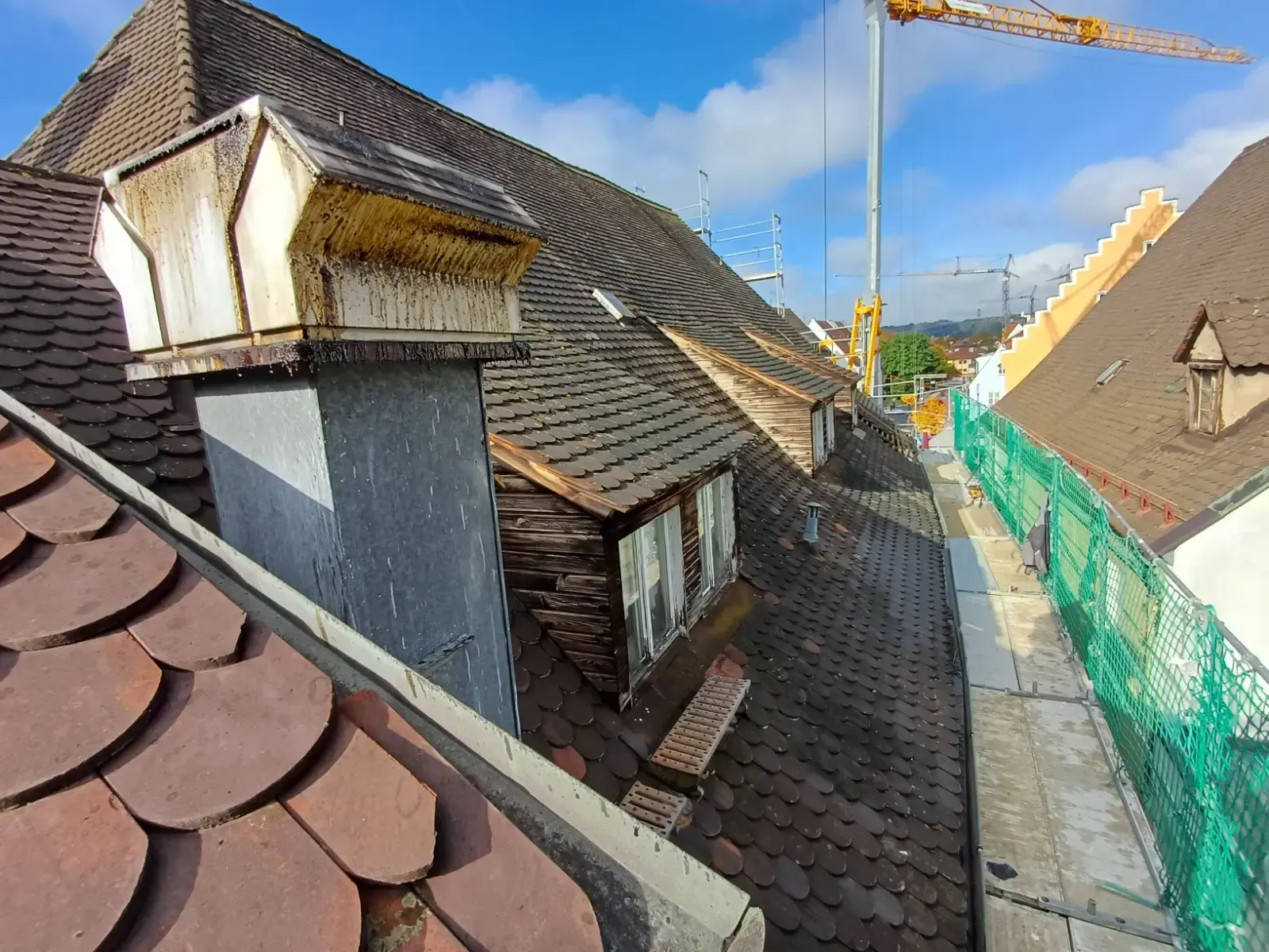 Blick vom Dach mit verwitterten Schindeln, Dachgauben und einem Kran unter blauem Himmel.