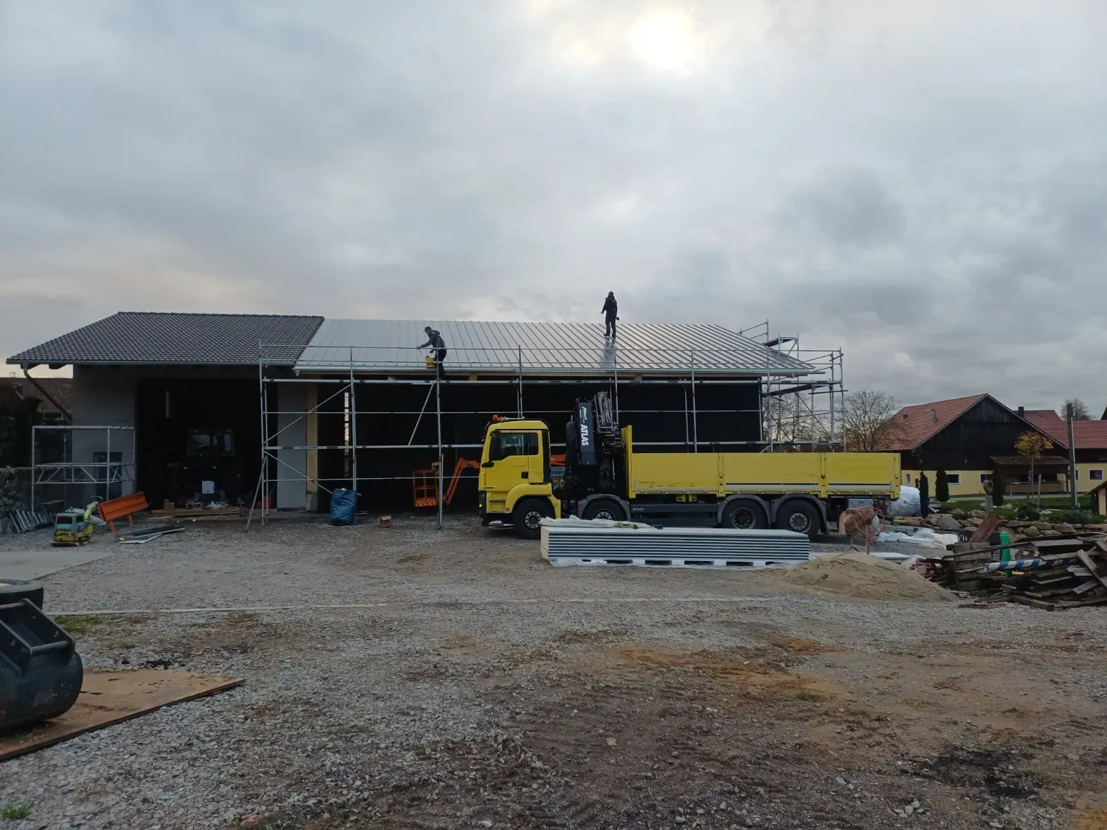 Baustelle mit gelbem Lastwagen, Arbeitern auf dem Dach und einem teilweise errichteten Haus unter bewölktem Himmel