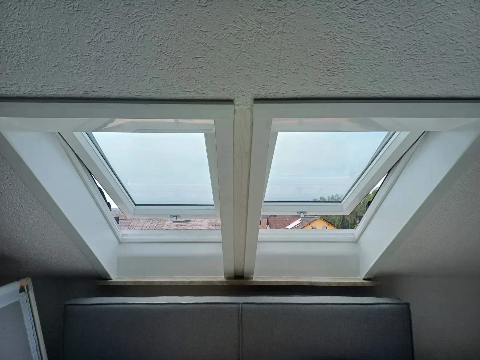 Zwei Dachfenster in einer weißen Decke über einem dunklen Sofa geben den Blick auf die Dächer draußen frei.