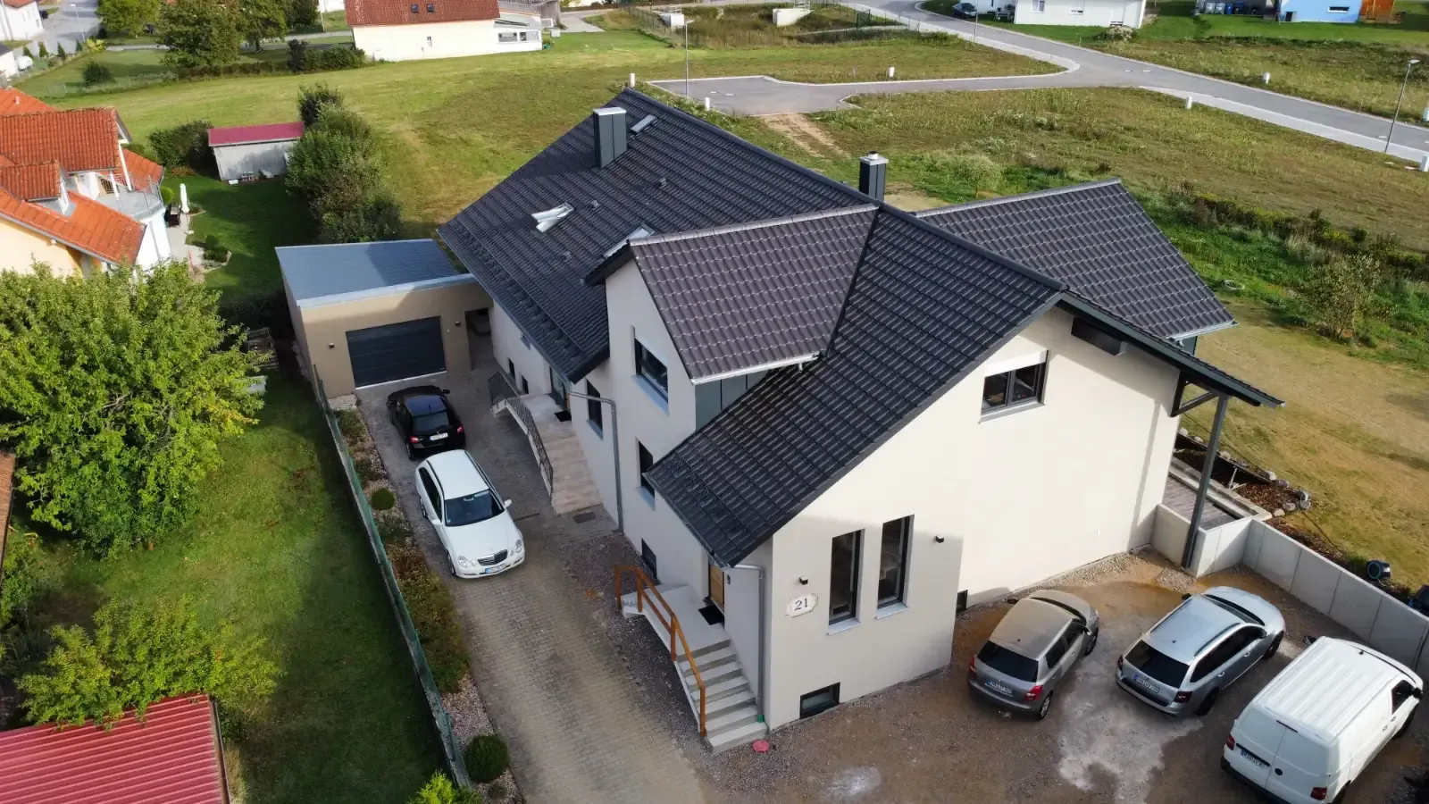 Luftaufnahme eines modernen weißen Hauses mit dunklem Dach, geparkten Autos und einer Einfahrt in einer ländlichen Gegend