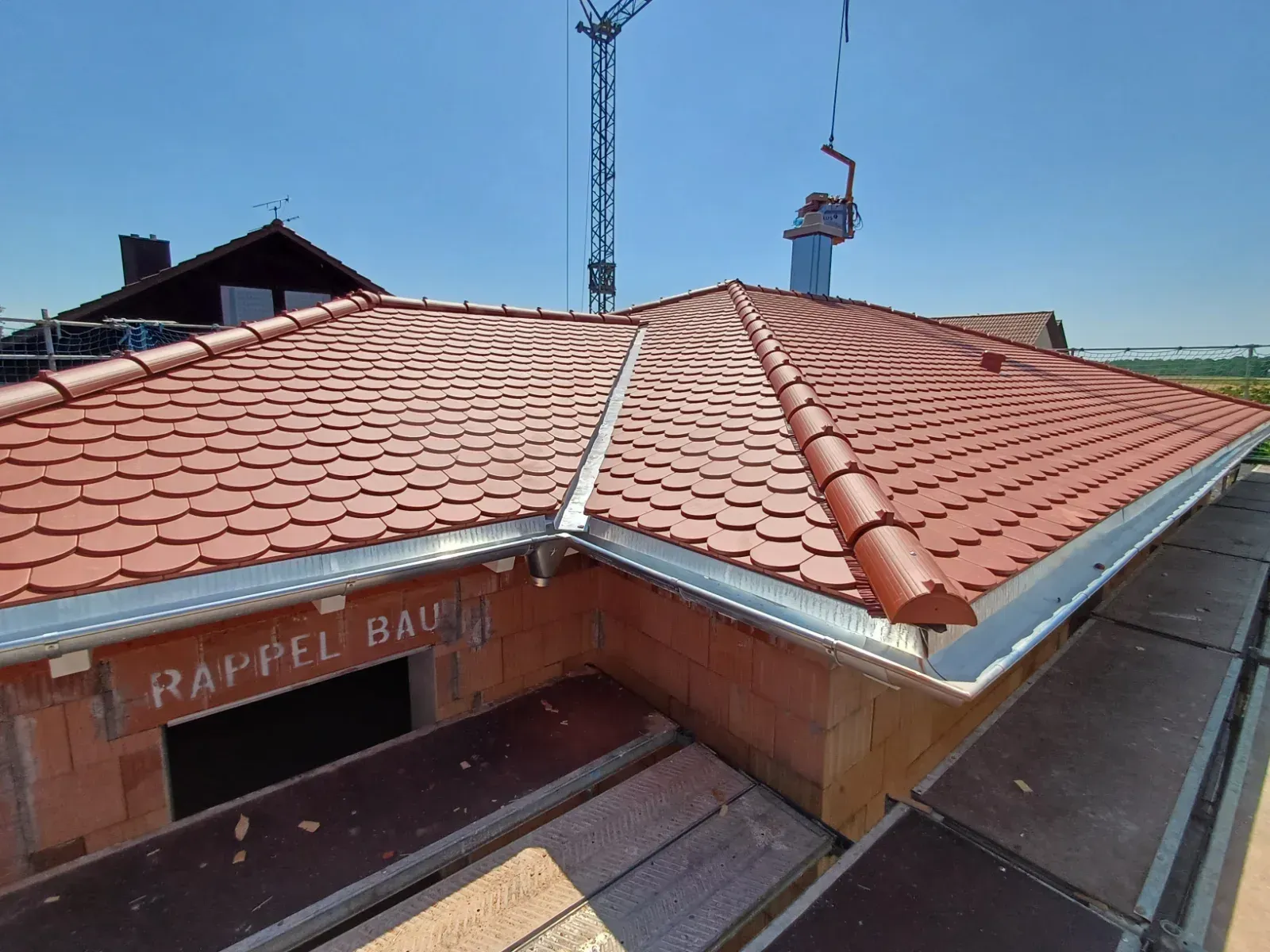 Ein im Bau befindliches Gebäude mit rotem Ziegeldach, im Hintergrund sind ein Schornstein und ein Kran zu sehen.