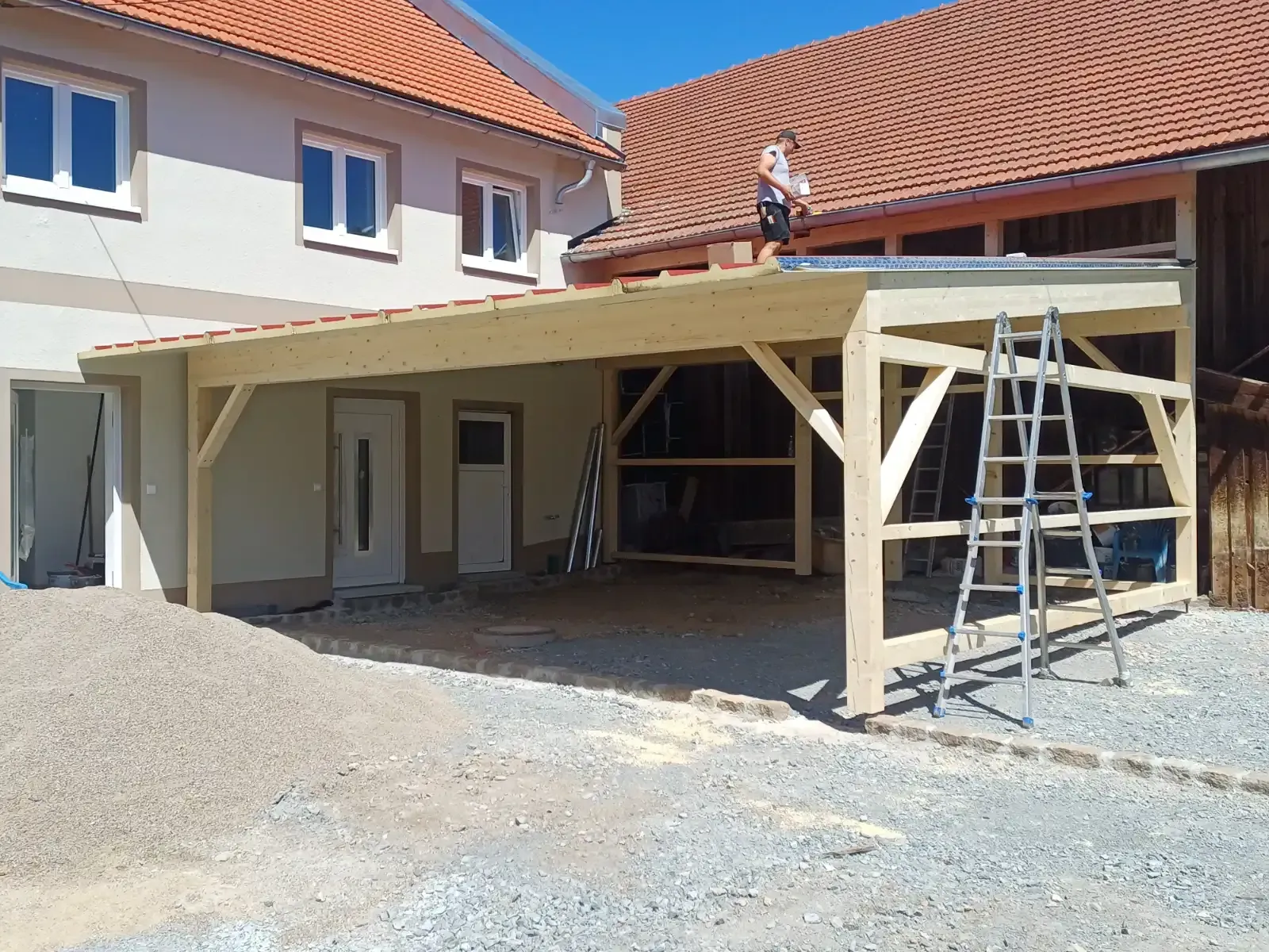 Neben einem Haus befindet sich ein großer, im Bau befindlicher Carport aus Holz; auf einer Kiesauffahrt stehen Leitern und Werkzeuge.