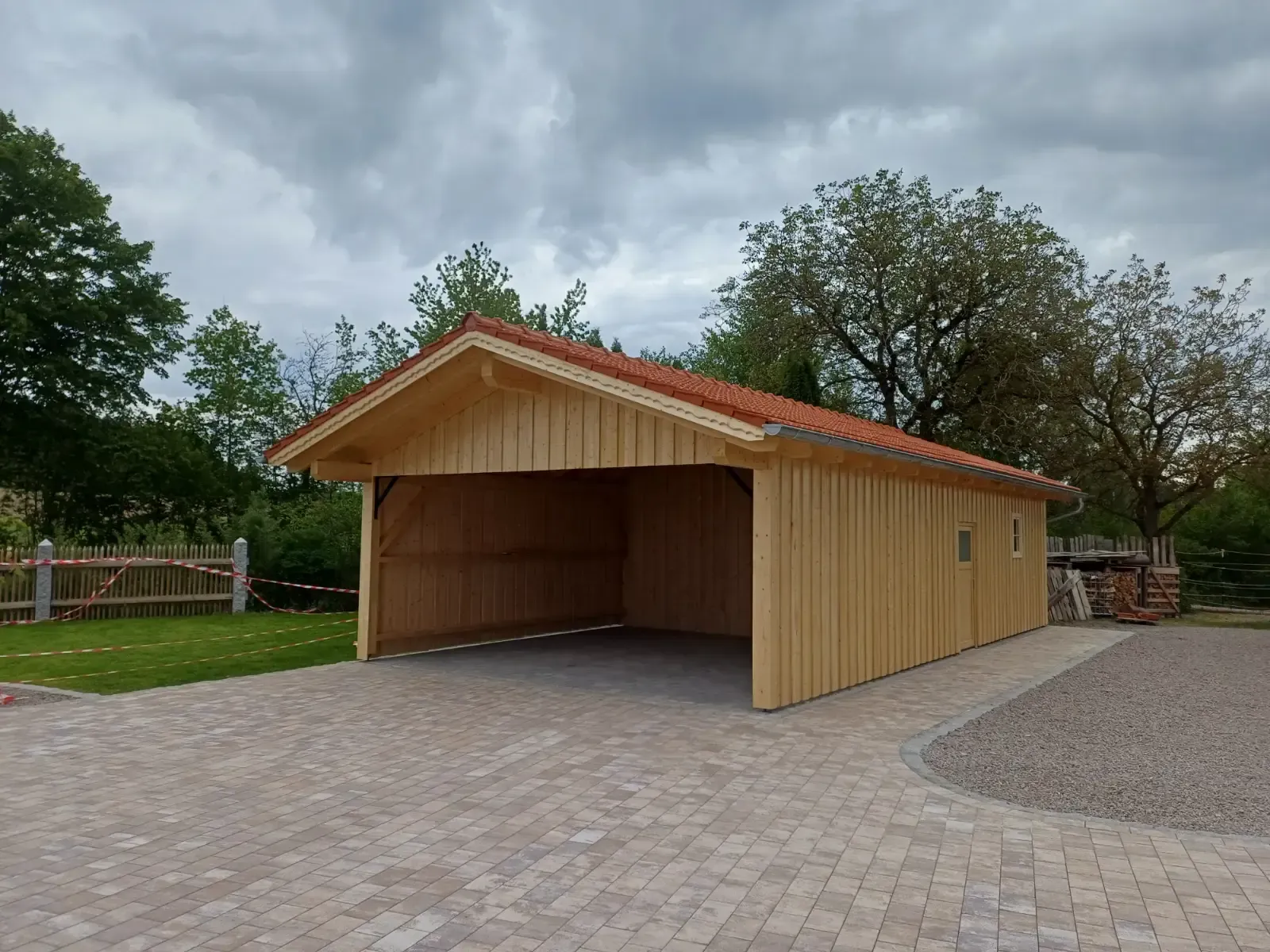 Ein offener Holzschuppen mit rotem Dach auf einem gepflasterten Platz, umgeben von Bäumen unter einem bewölkten Himmel