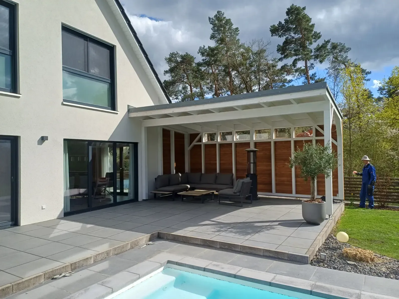 Moderne Hausterrasse mit weißer Pergola, Gartensofa, Steinpflaster und einer Person, die am Rasen steht