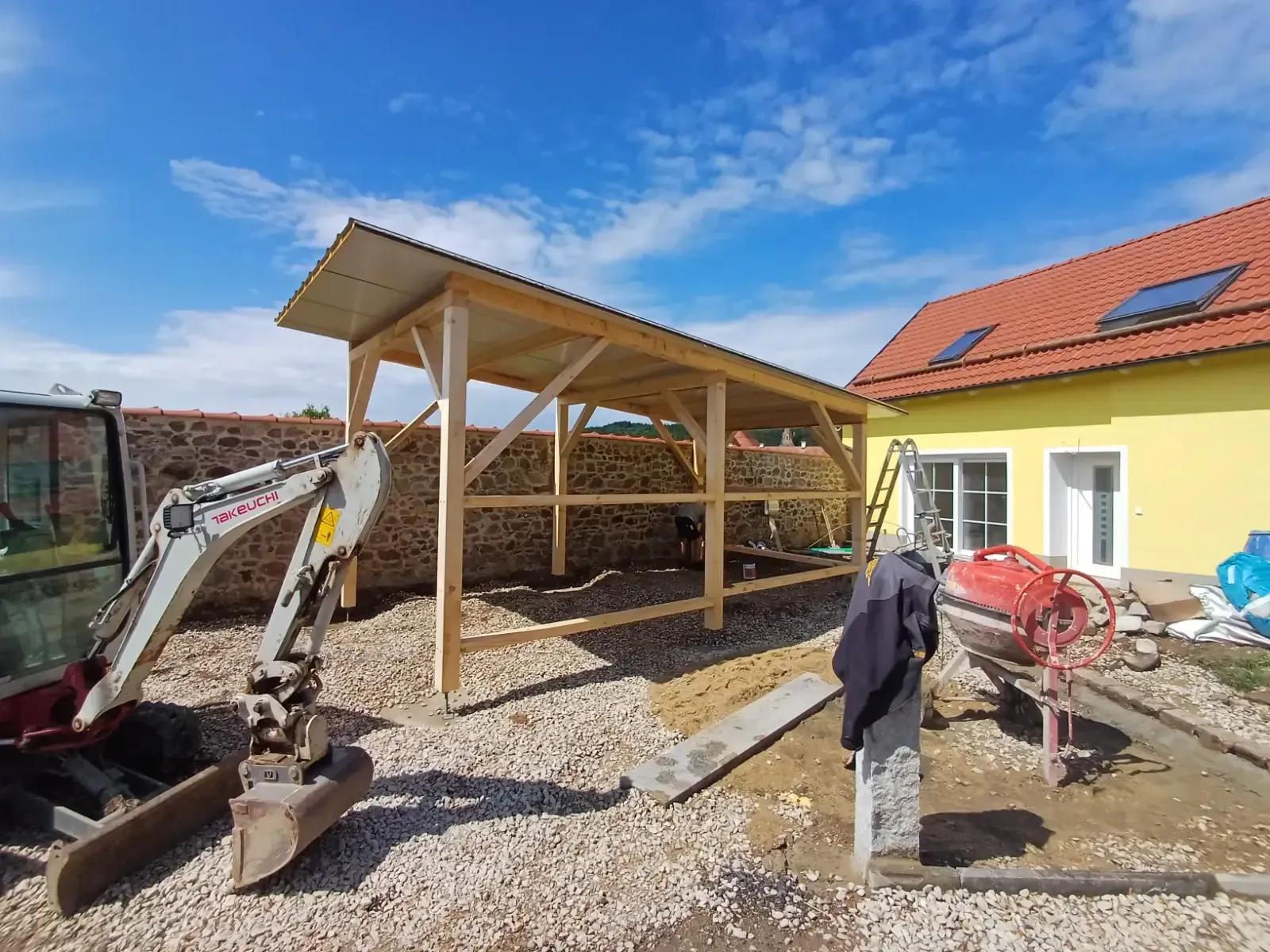 Baustelle mit hölzernem Carportgerüst, Kiesboden, Bagger und gelbem Haus unter blauem Himmel