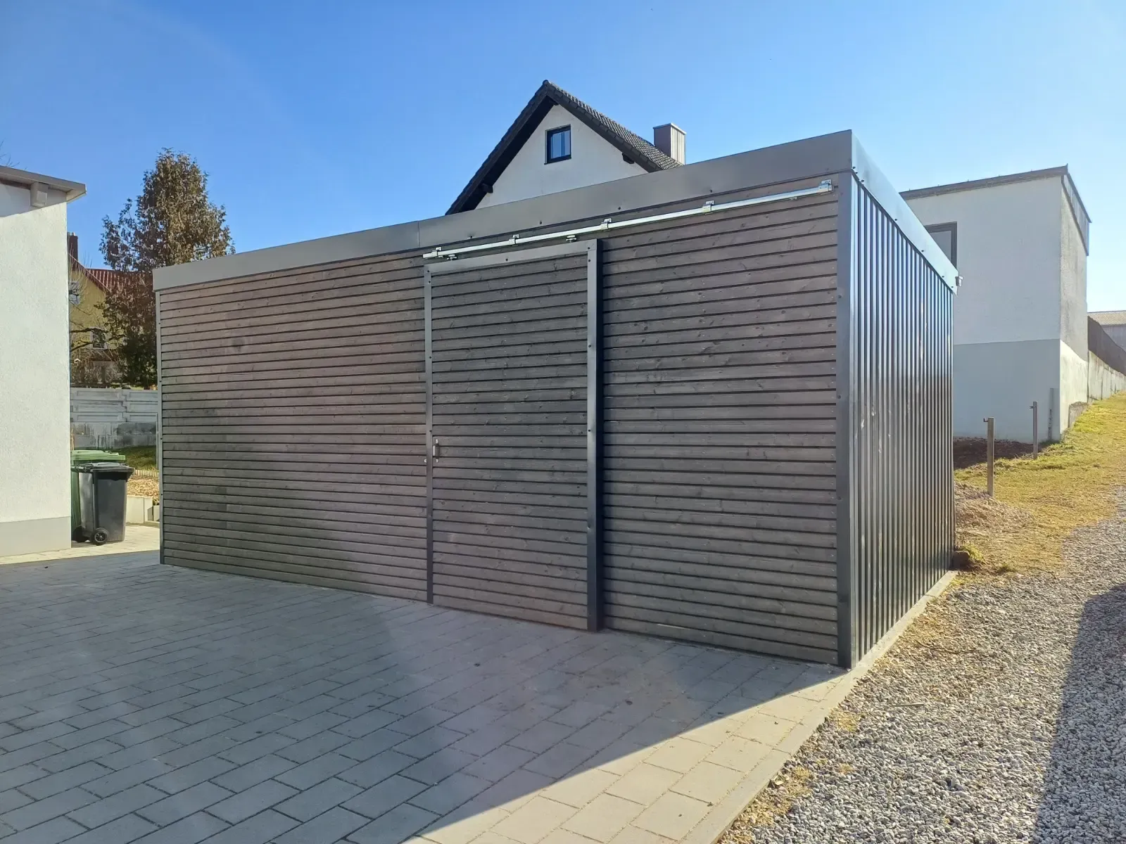 Moderne, graue Garage oder Lagerhalle mit Lattenverkleidung neben einer gepflasterten Auffahrt und einem Kieshof
