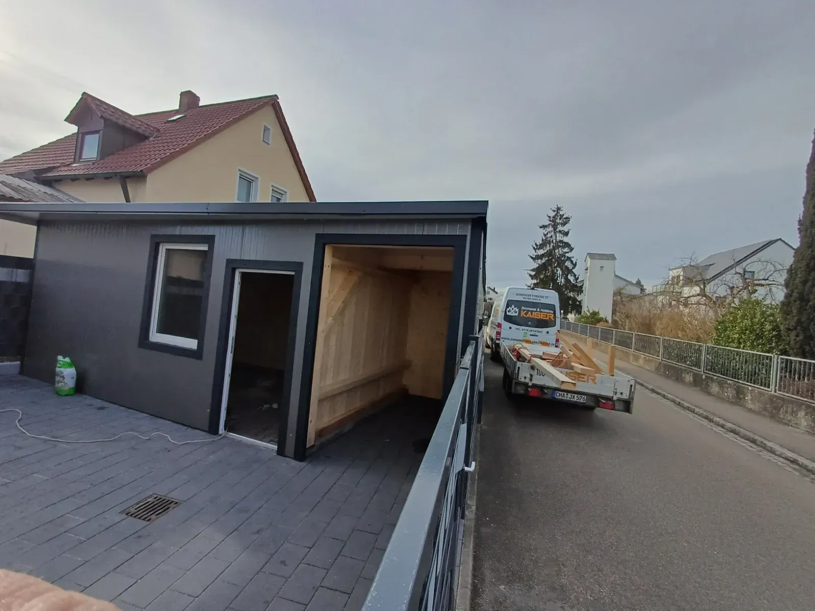 Schwarzer Gebäudeanbau neben einer Straße, auf der ein kleiner Anhänger geparkt ist.
