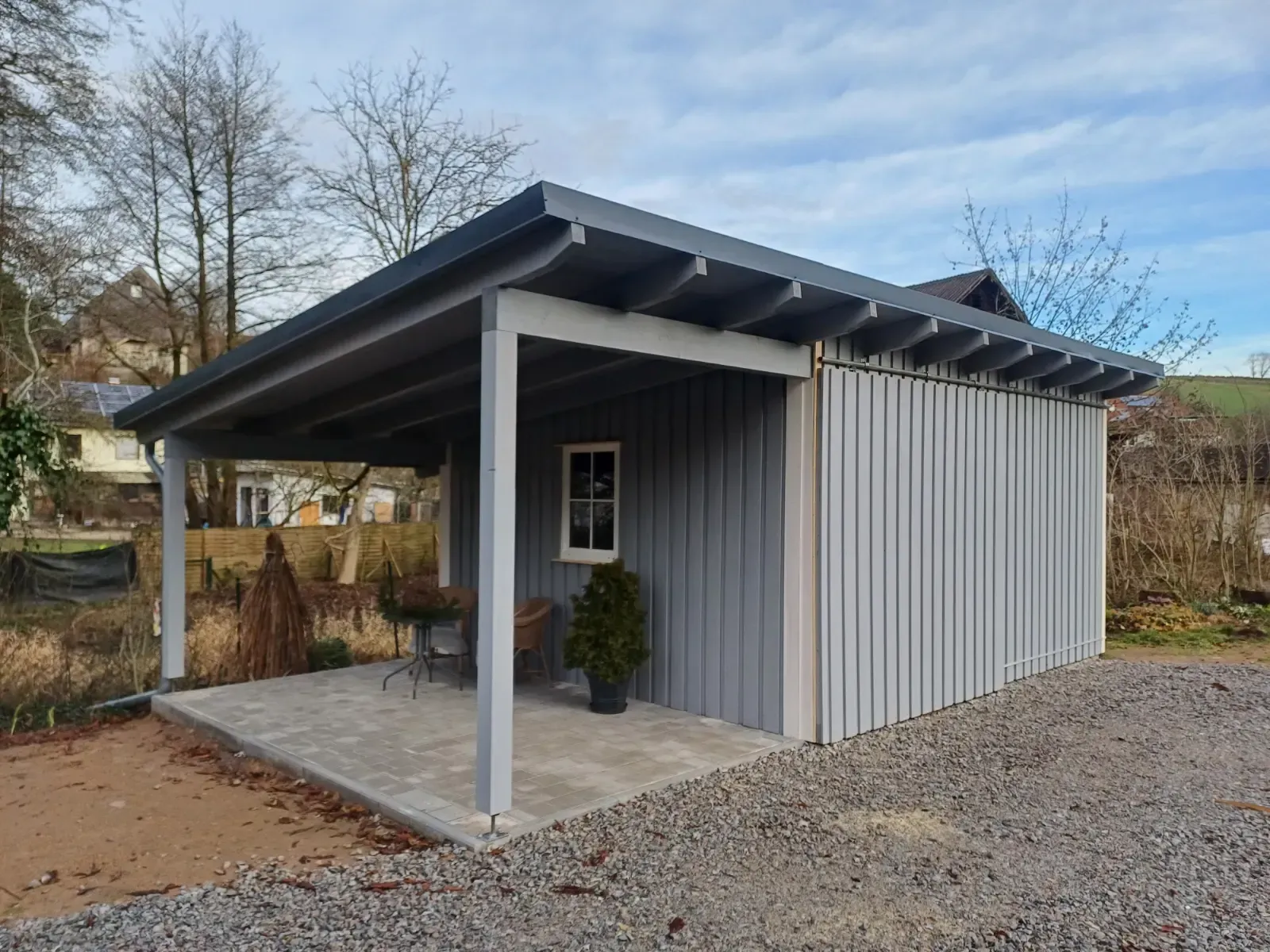 Kleiner grauer Gartenschuppen mit überdachter Veranda und schrägem Dach auf einem Kieshof.