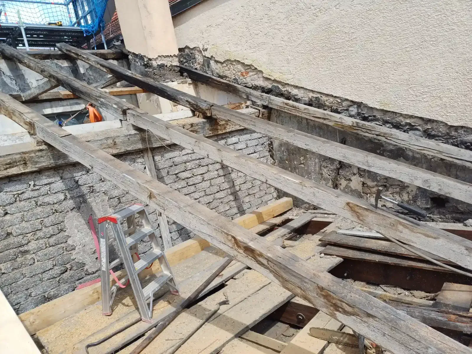Eingestürzte hölzerne Dachbalken und Trümmer neben einer Mauer, dazwischen eine Metallleiter.