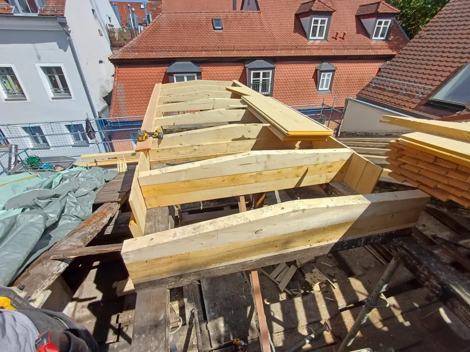 Auf einem Dach neben rot gedeckten Gebäuden werden hölzerne Dachstühle errichtet.