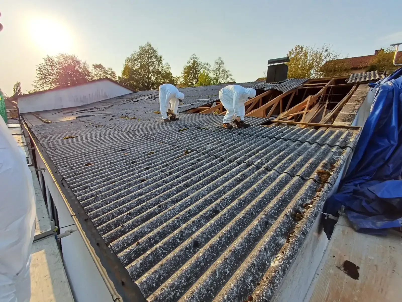 Arbeiter in weißen Schutzanzügen begutachten bei Sonnenuntergang ein Wellblechdach, das sich in Reparatur befindet.