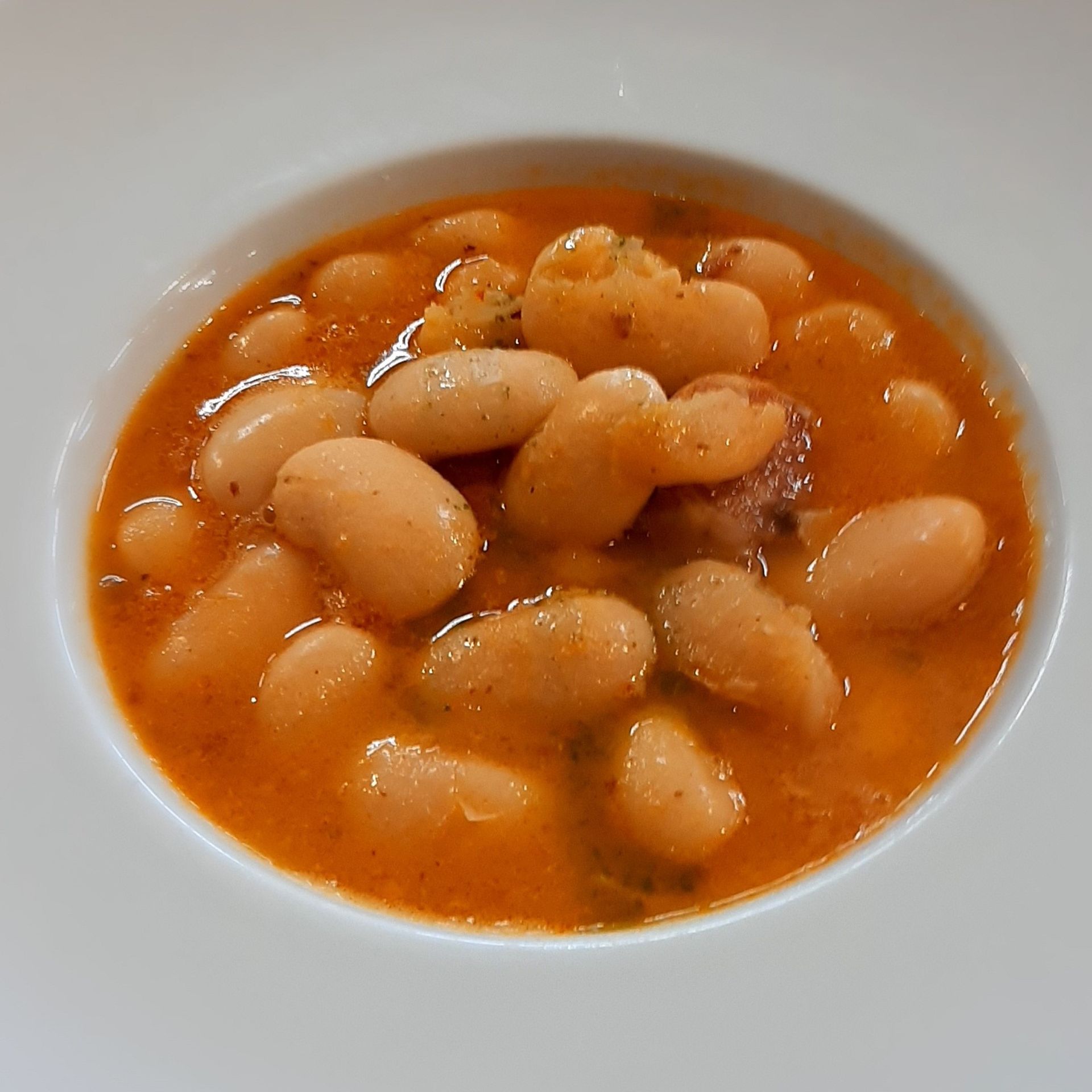 Un tazón blanco lleno de un guiso caliente a base de tomate con grandes alubias blancas y trozos de salchicha.