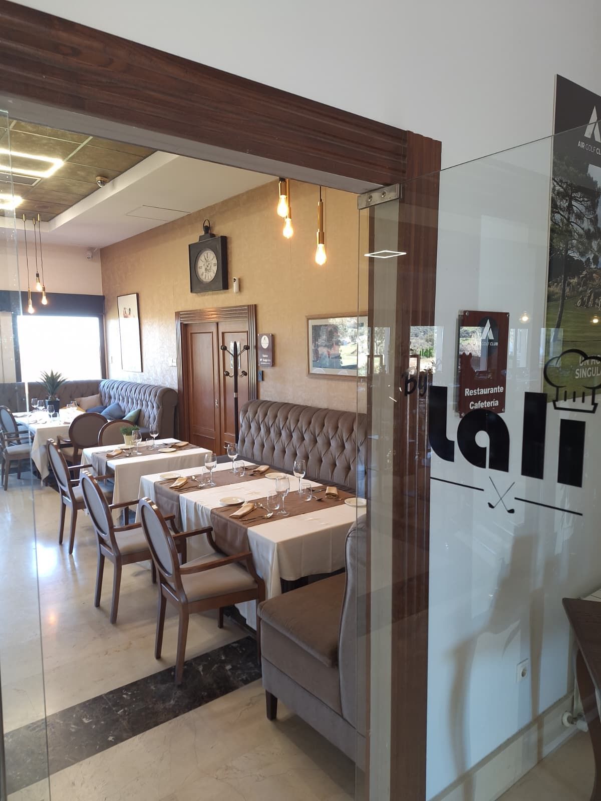 Una vista a través de una puerta de cristal hacia un restaurante bien iluminado con asientos tipo cabina tapizados, manteles blancos y relojes de pared.