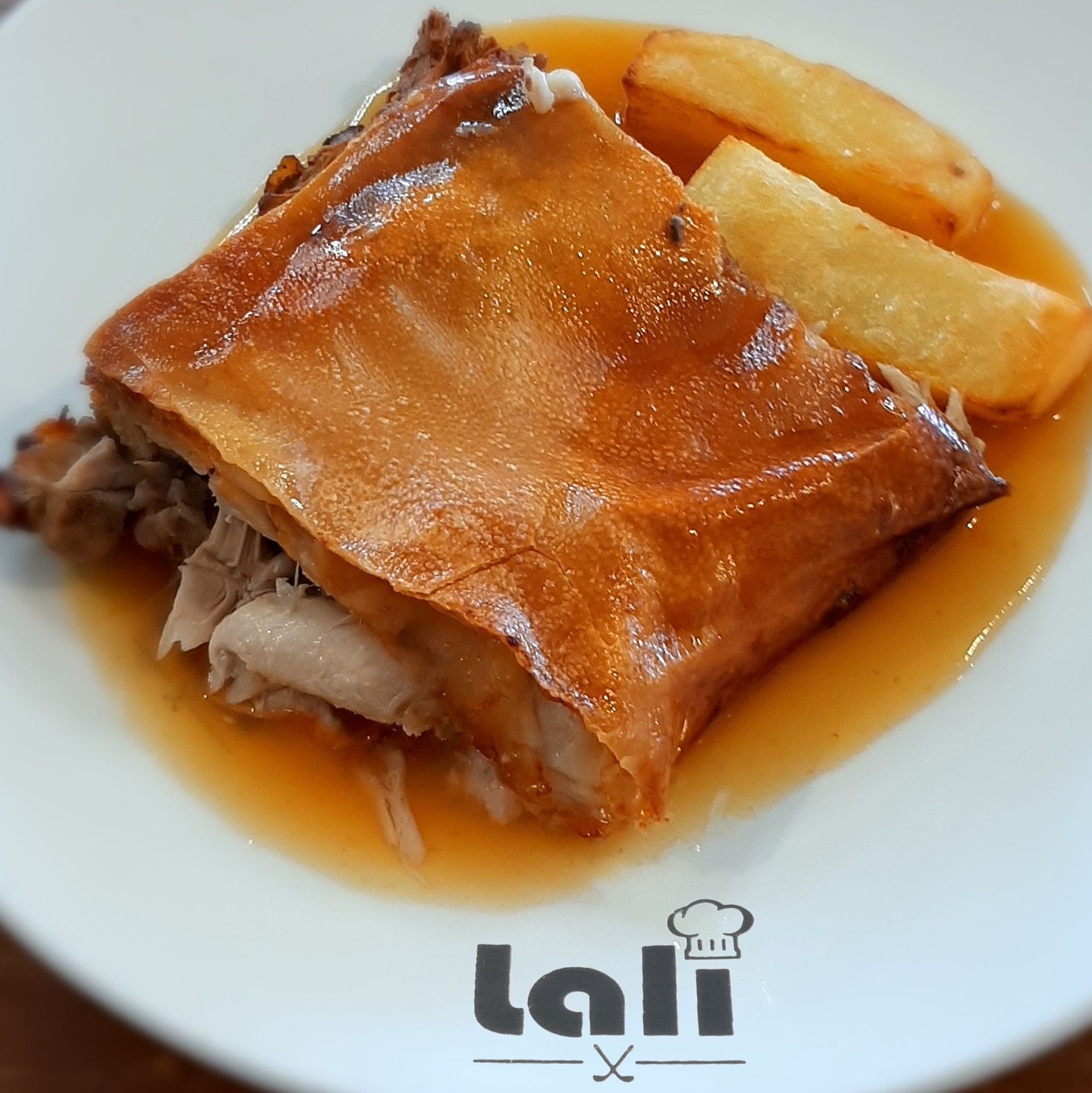 Un plato de cerdo asado con piel crujiente y patatas fritas, servido en un glaseado sabroso sobre un plato blanco con el logotipo del restaurante.
