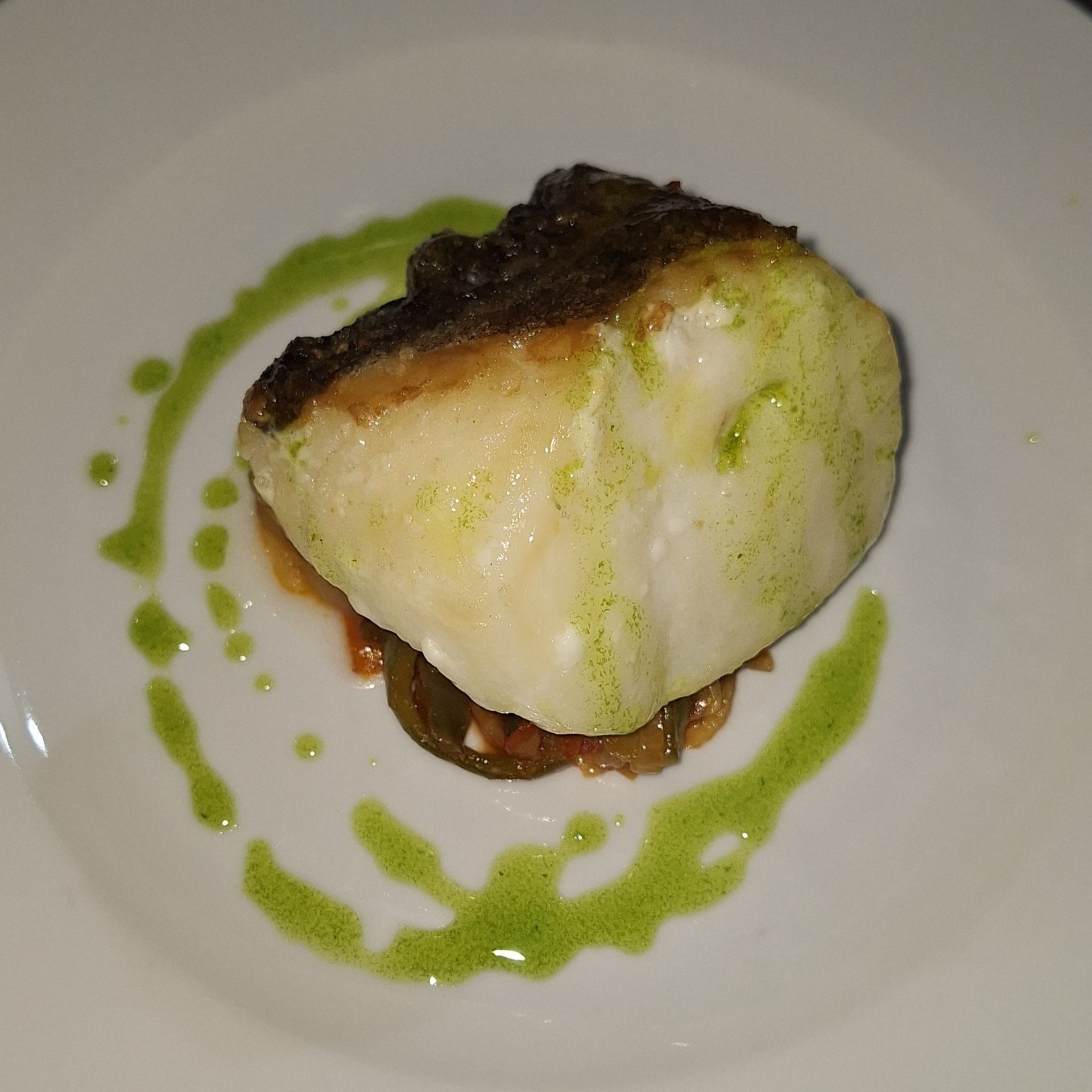 Un filete de pescado sellado, servido sobre una guarnición de verduras en un plato blanco y adornado con un chorrito circular de aceite verde.