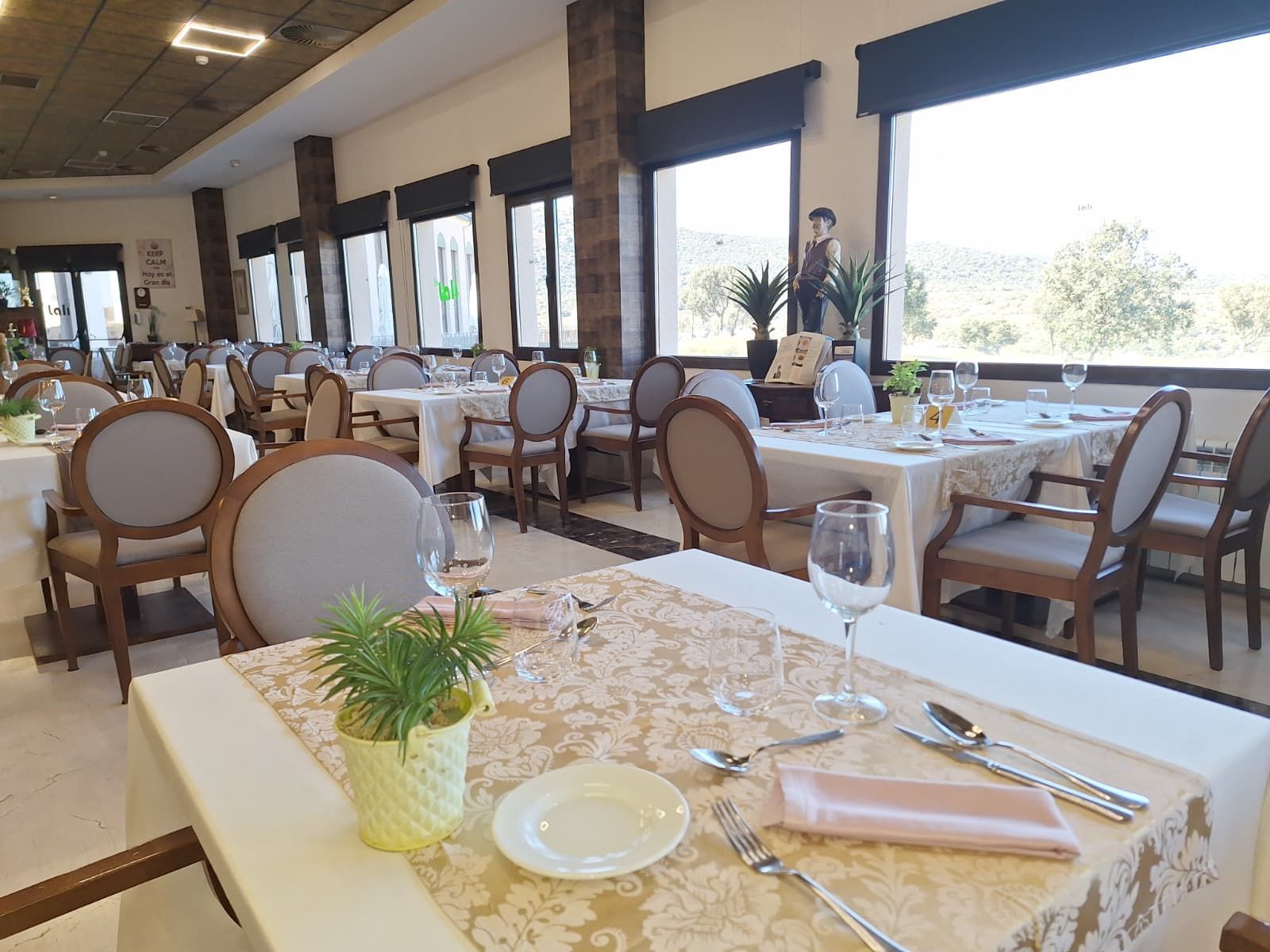 Un comedor de restaurante con filas de mesas con manteles blancos, caminos de mesa florales y vajilla puesta cerca de grandes ventanales.