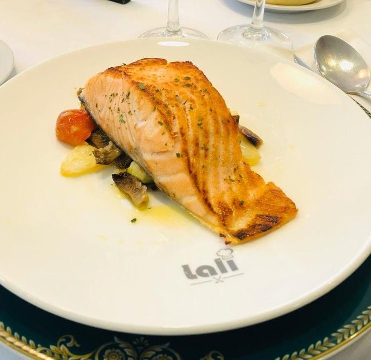 Un filete de salmón sellado, servido sobre una cama de patatas y champiñones asados ​​con un tomate cherry, en un plato de la marca Lali.