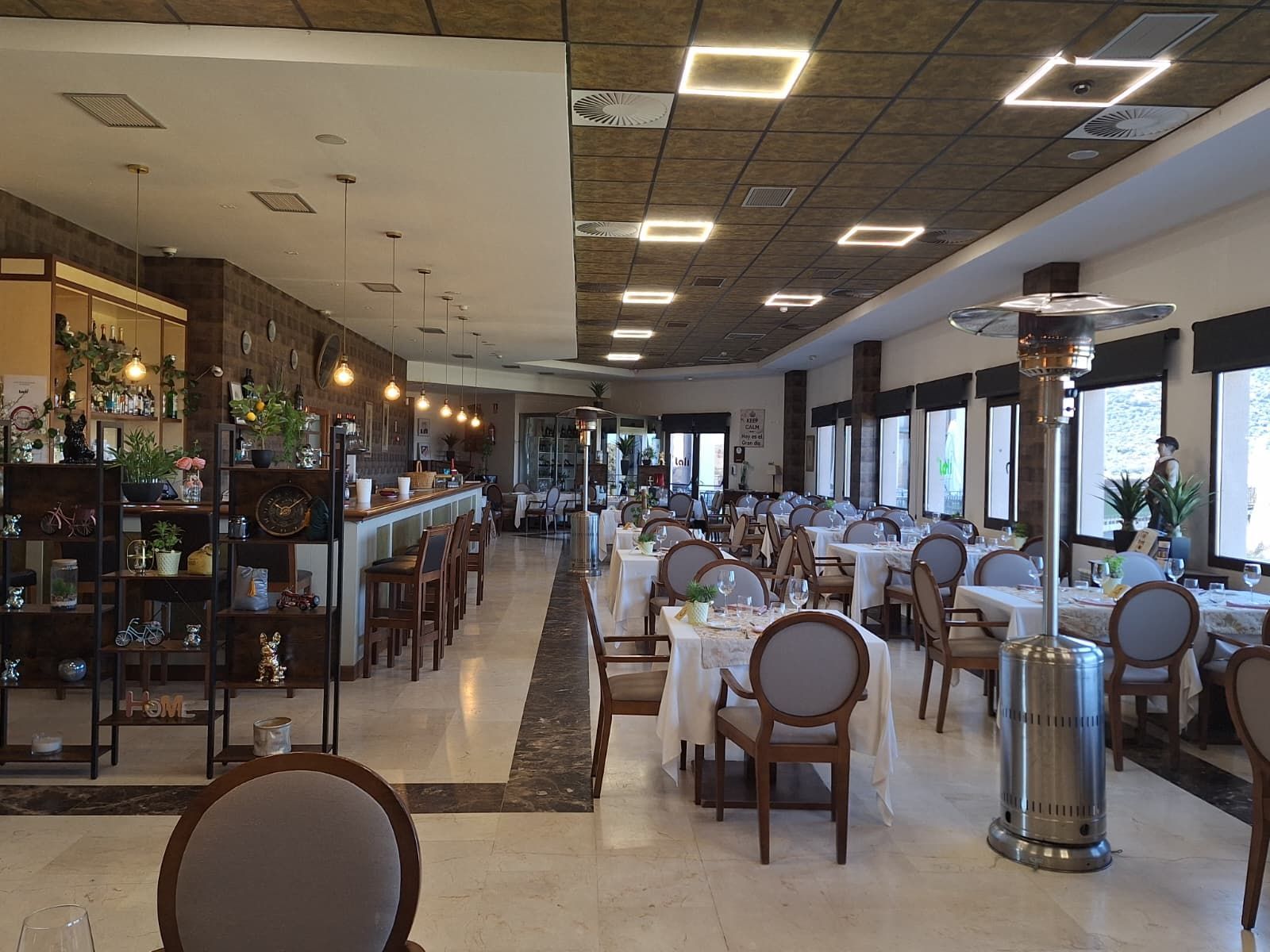 El interior de un comedor con una barra a la izquierda, filas de mesas puestas con sillas grises y un calefactor de patio alto en el centro.