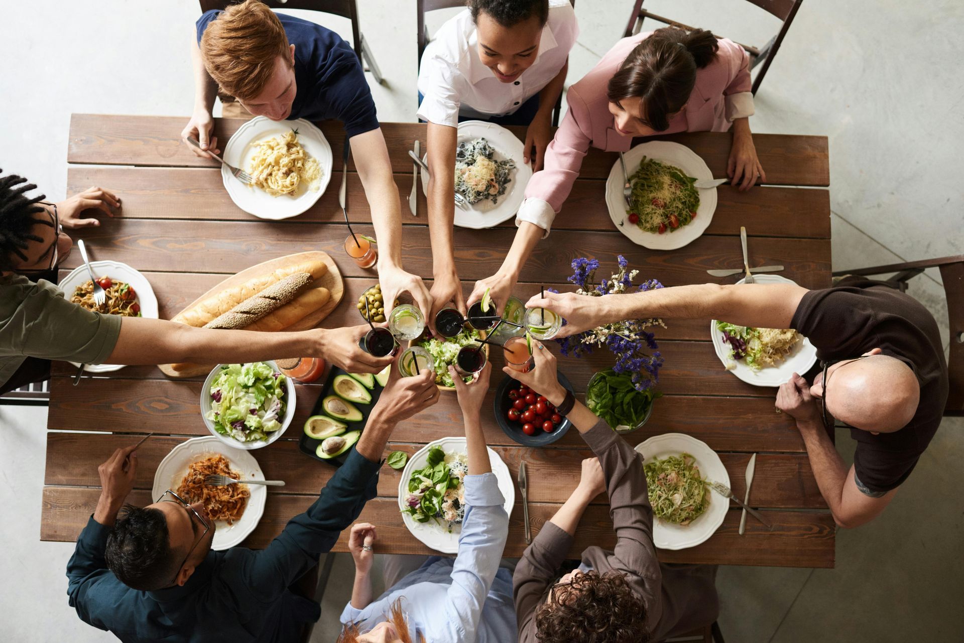Un grupo de personas sentadas alrededor de una mesa de madera, brindando con vasos mientras disfrutan de una comida de pasta, pan y ensalada.