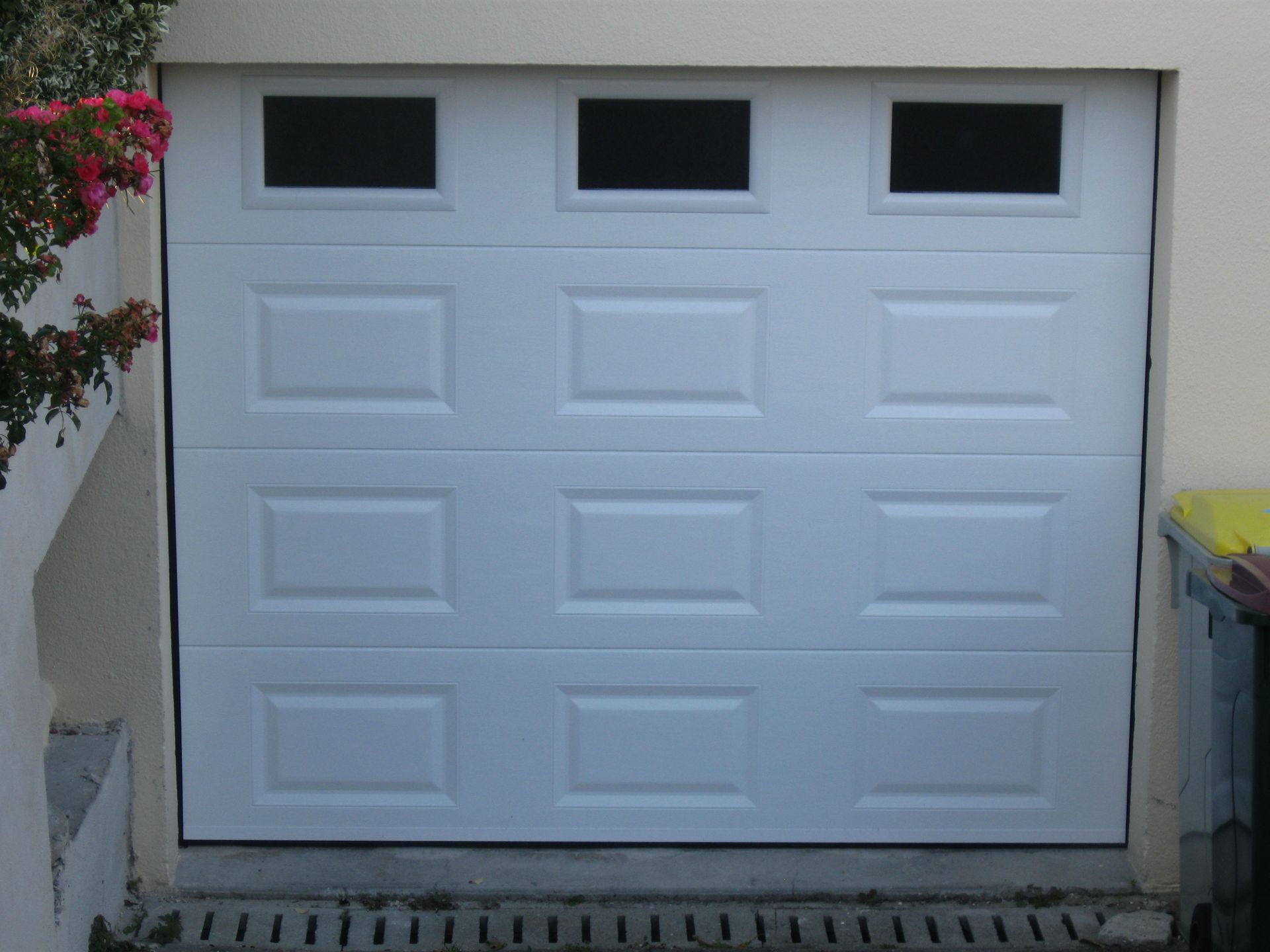 Porte de garage blanche avec trois petites fenêtres en partie supérieure, encastrée dans un mur extérieur en stuc.