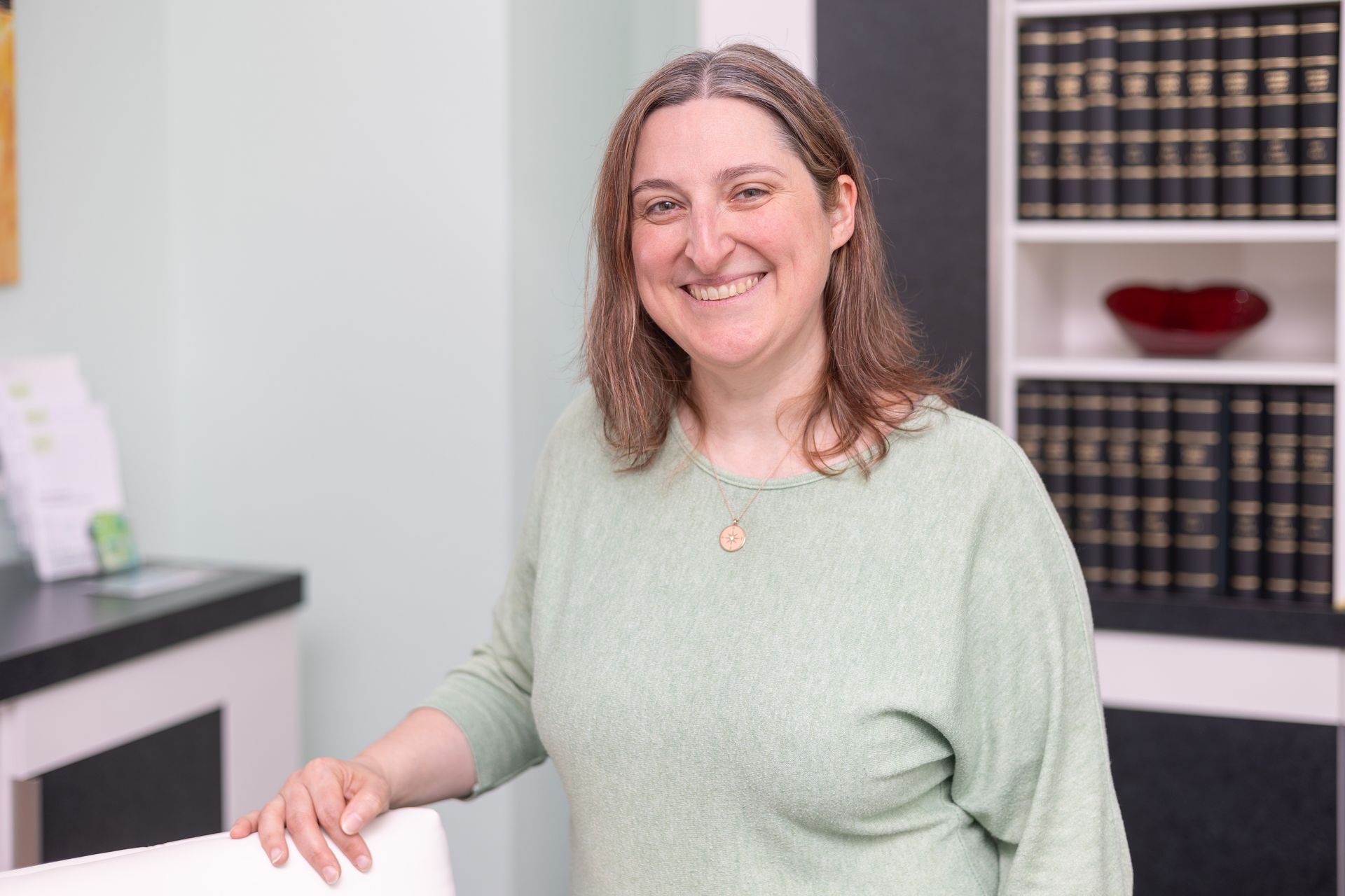 Eine Frau in einem grünen Pullover steht vor einem Bücherregal und lächelt.