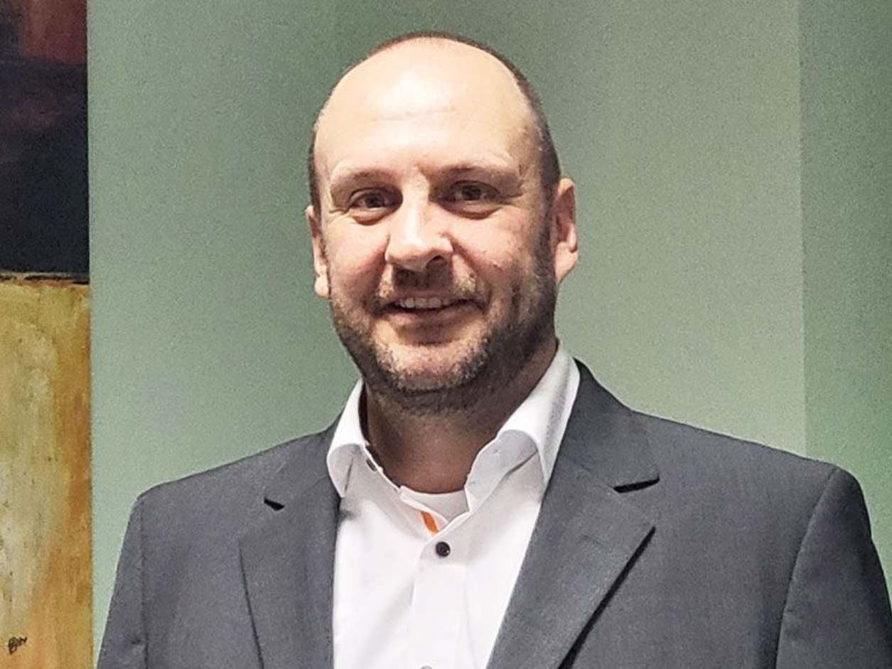 Ein Mann in Anzug und weißem Hemd steht vor einem Bücherregal.