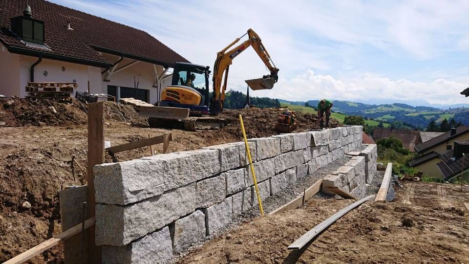 Ein Bagger arbeitet an einer Steinmauer vor einem Haus.