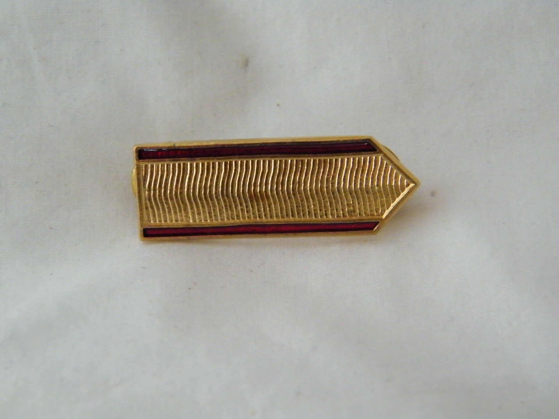 Un alfiler dorado con una raya roja reposa sobre una superficie blanca.