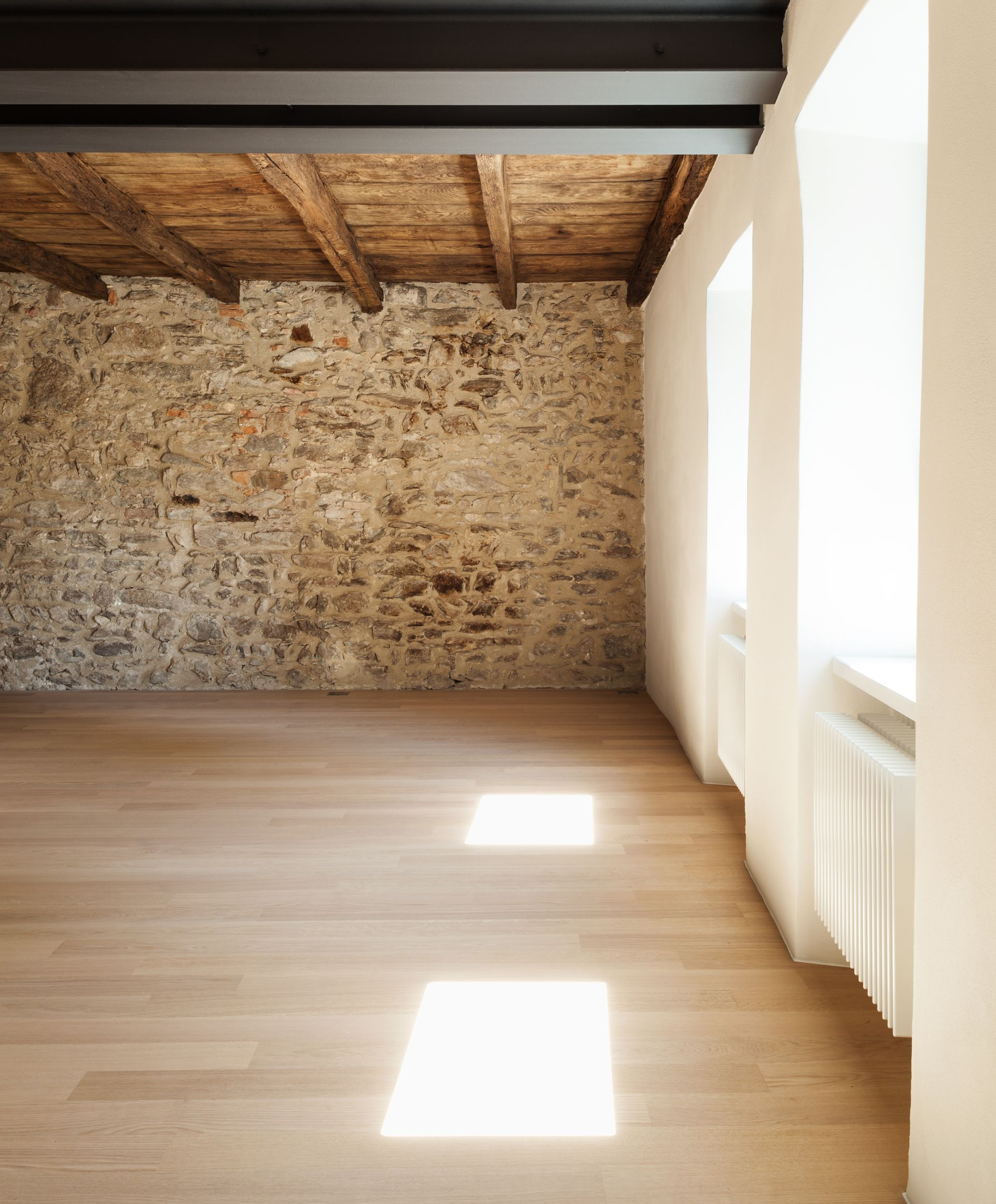 Pièce vide avec un mur en pierre, un plafond à poutres apparentes, un parquet en chêne clair et des murs blancs.