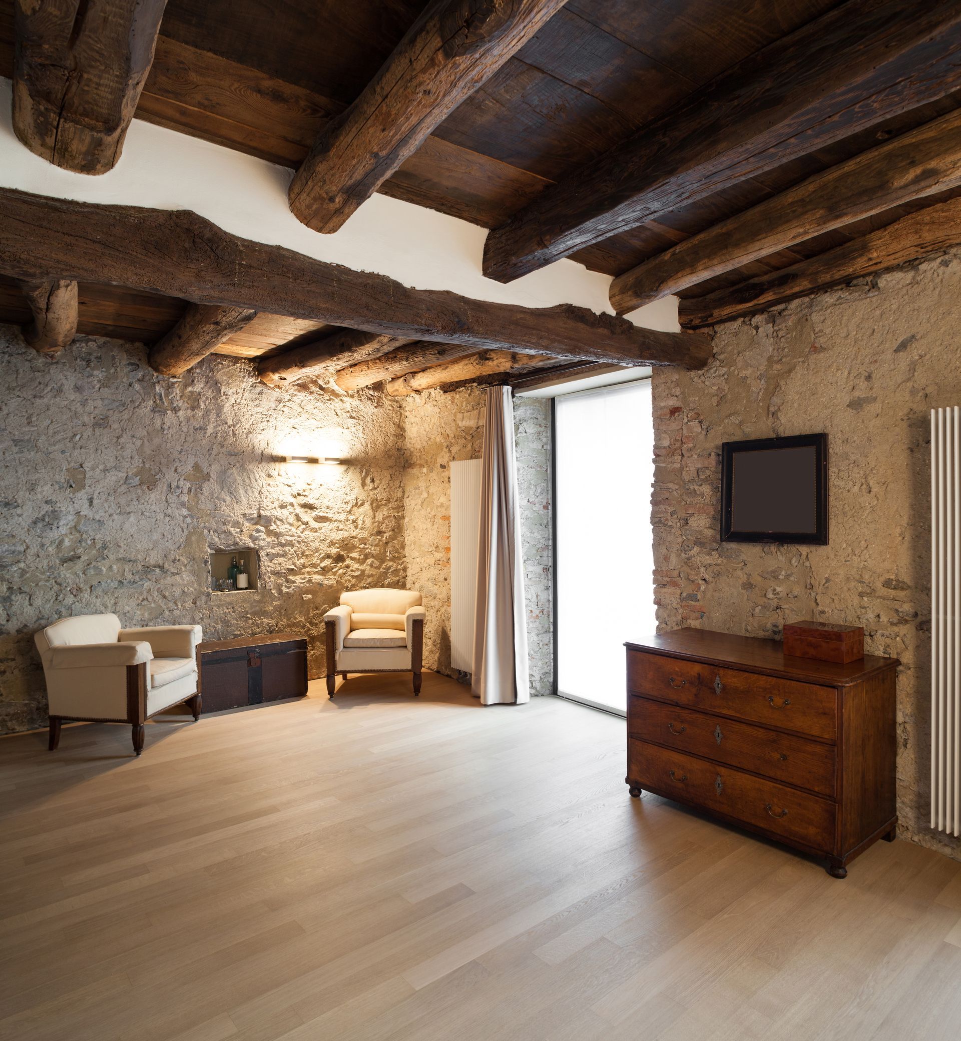Une pièce rustique avec des murs en pierre, des poutres au plafond, un parquet clair, deux fauteuils et une commode en bois.