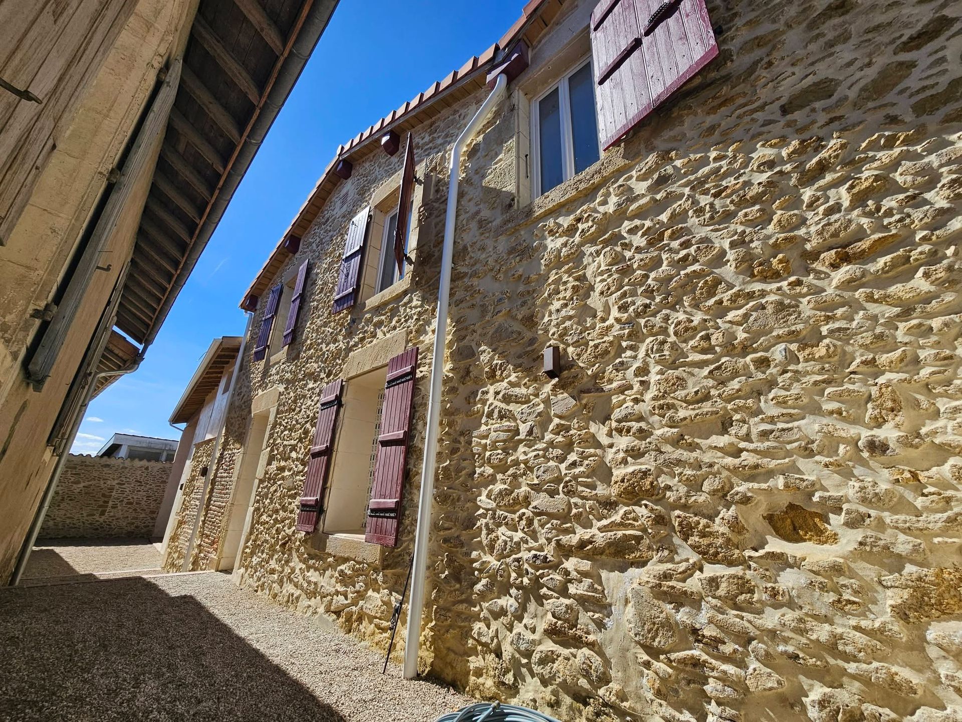 Vue en contre-plongée d'un bâtiment rustique en pierre aux volets bordeaux, sous un ciel d'un bleu éclatant.