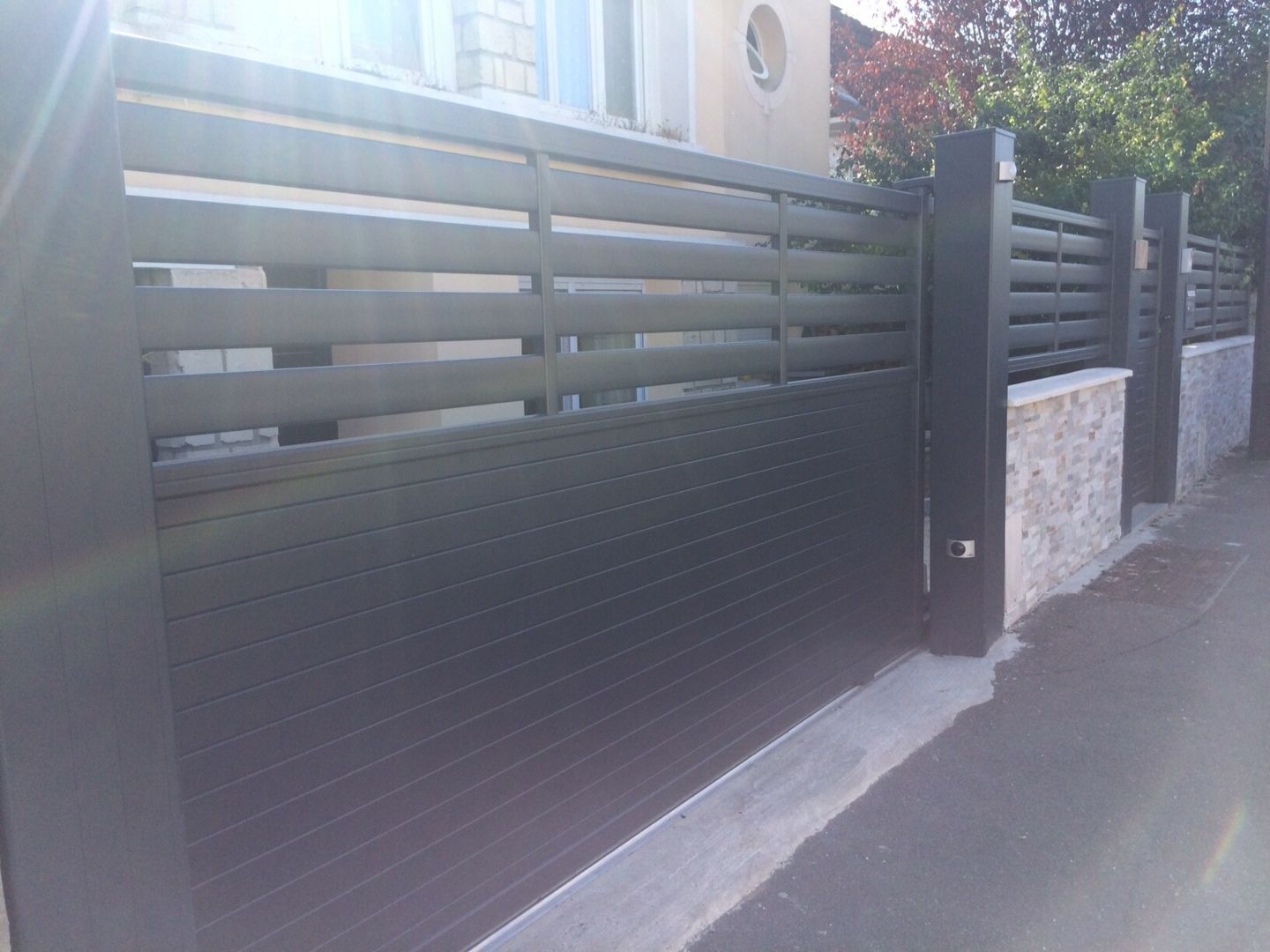 Une clôture moderne à lattes horizontales gris foncé avec une base en piliers de pierre borde une rue pavée.