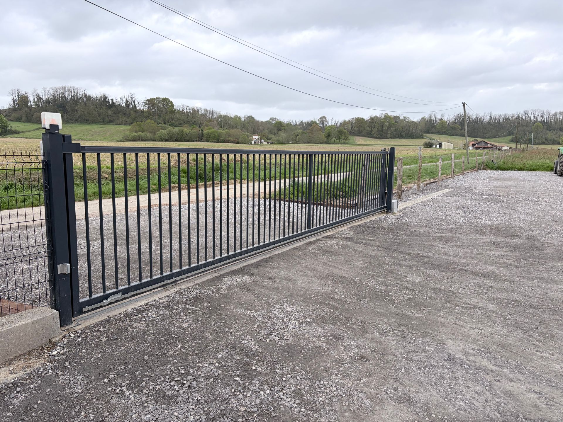 Un portail en métal noir barre une allée de gravier, avec en arrière-plan un champ et un ciel nuageux.