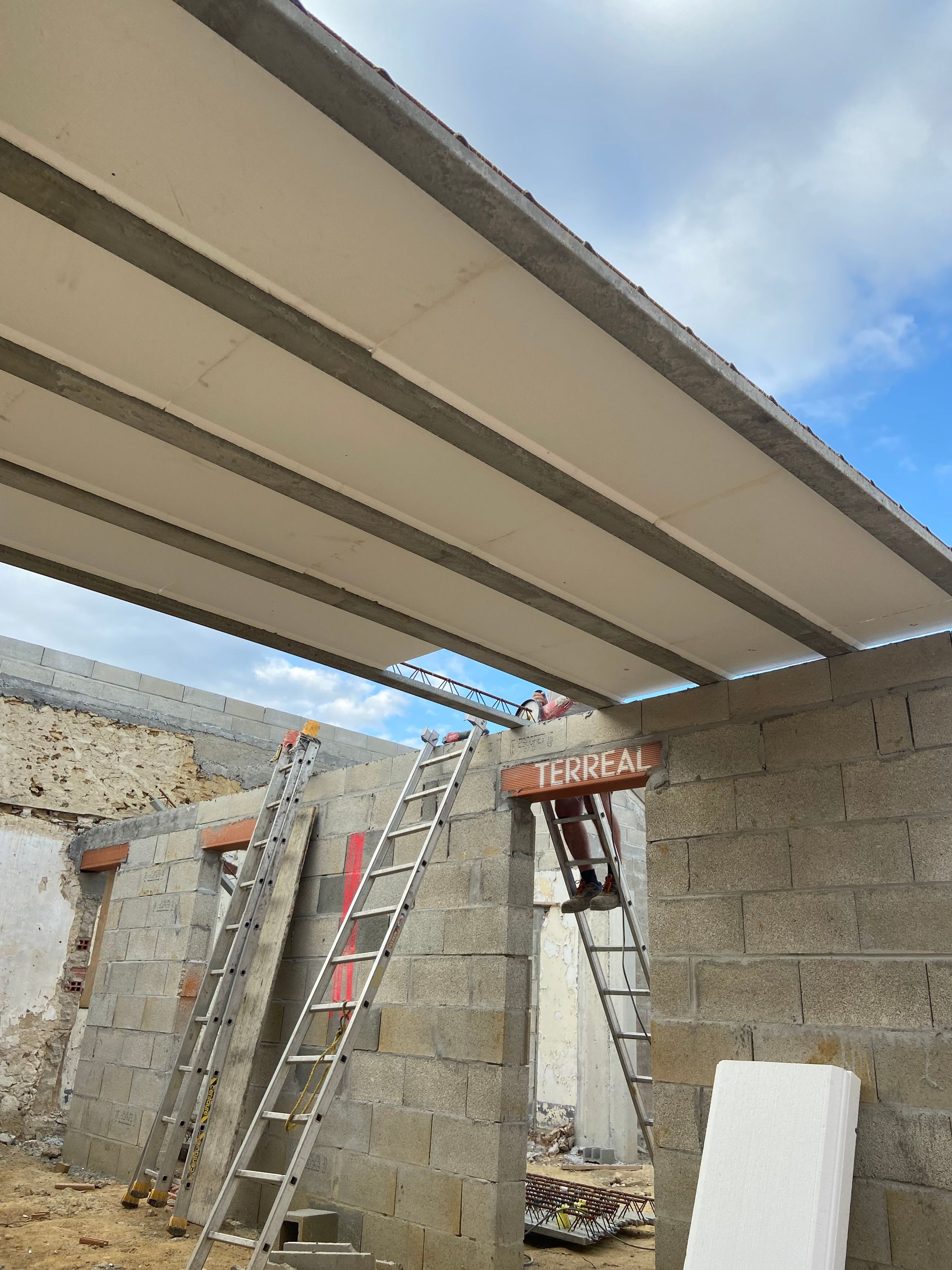 Chantier de construction montrant des murs en blocs de béton gris, des panneaux de toiture métalliques et deux échelles.