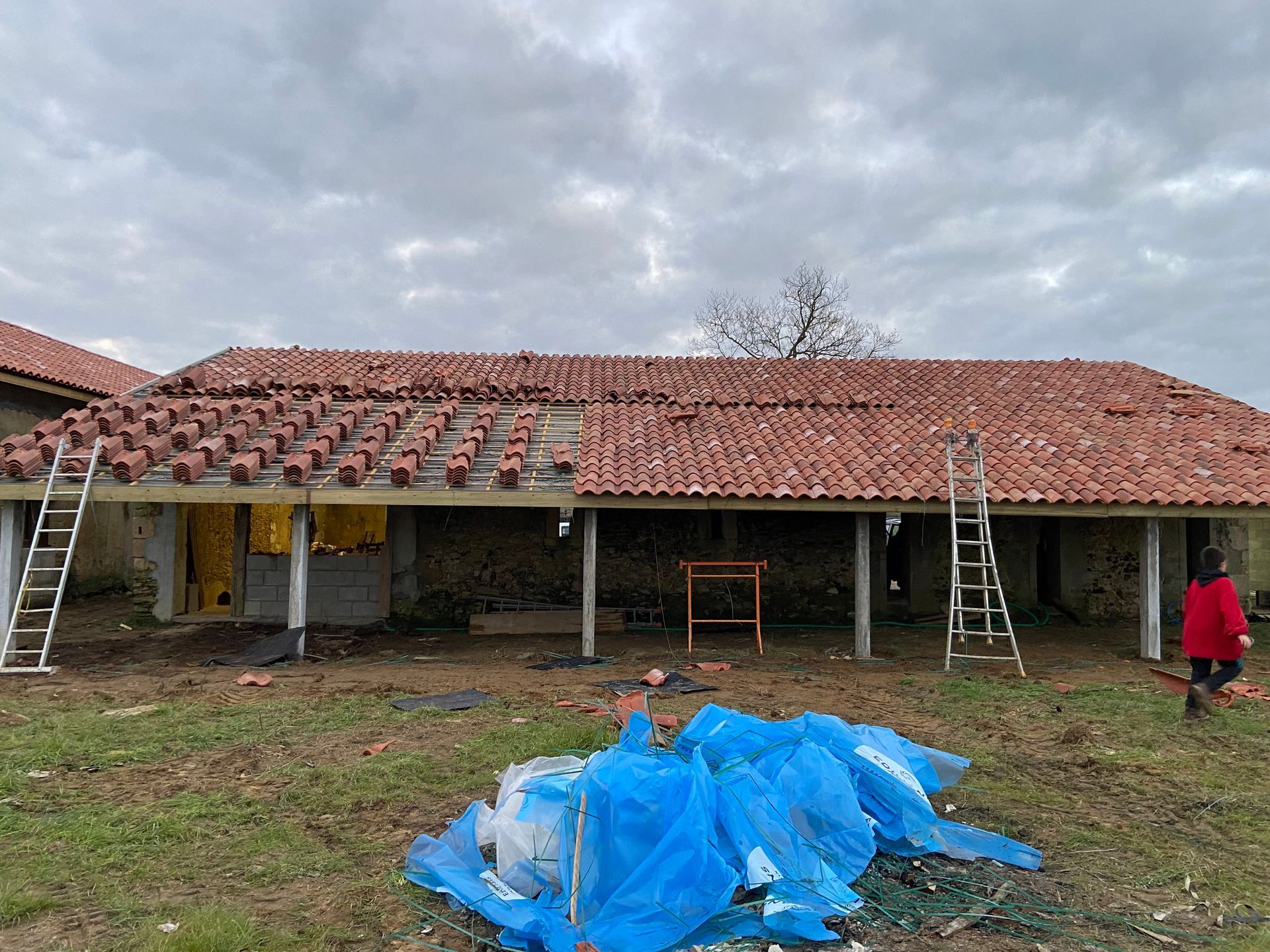 Une personne se tient près d'un bâtiment en rénovation, où une partie du toit en tuiles de terre cuite est réparée.