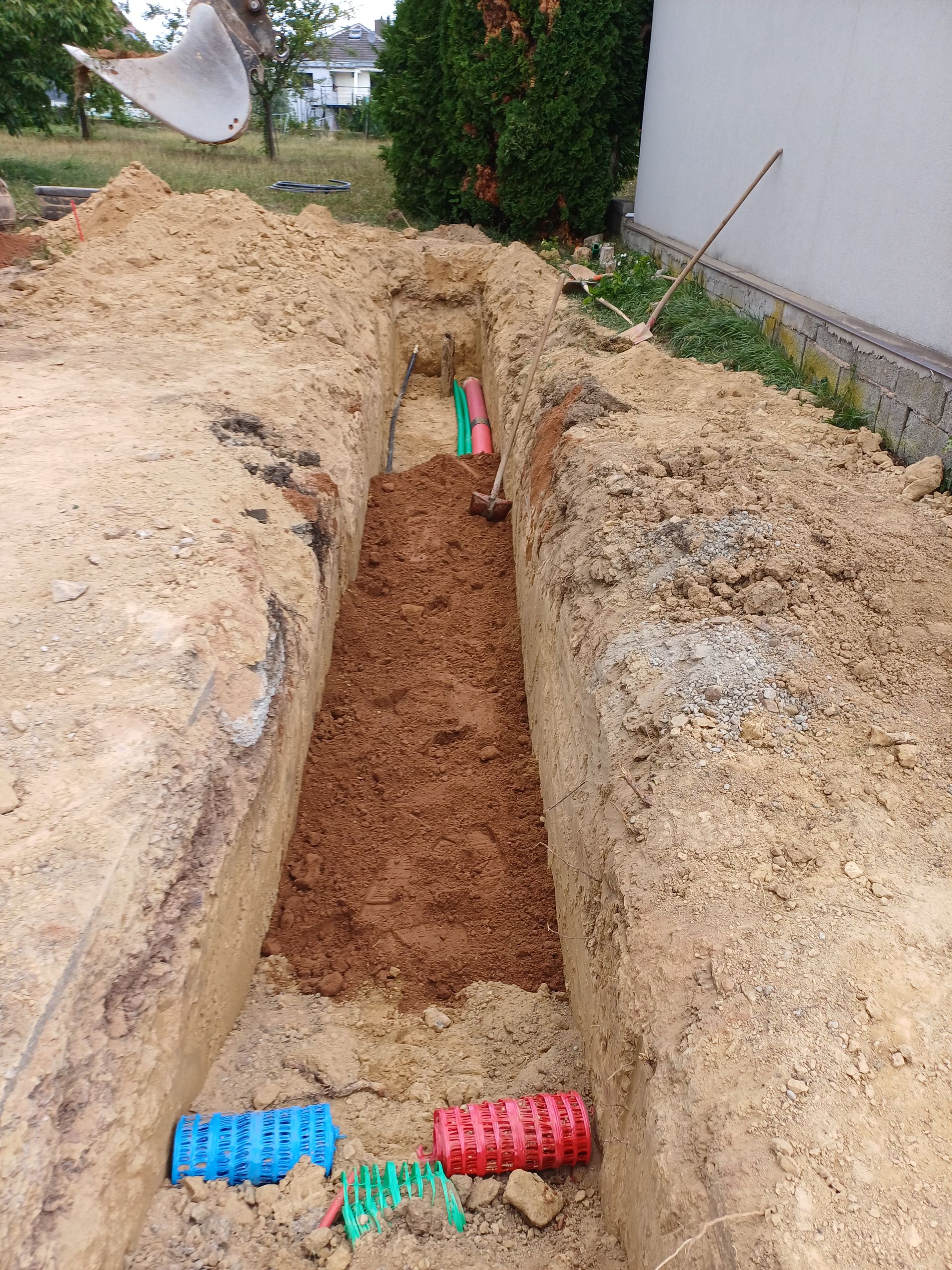 Tranchée avec tuyaux colorés pour les services publics, creusée dans la terre près d'un bâtiment.