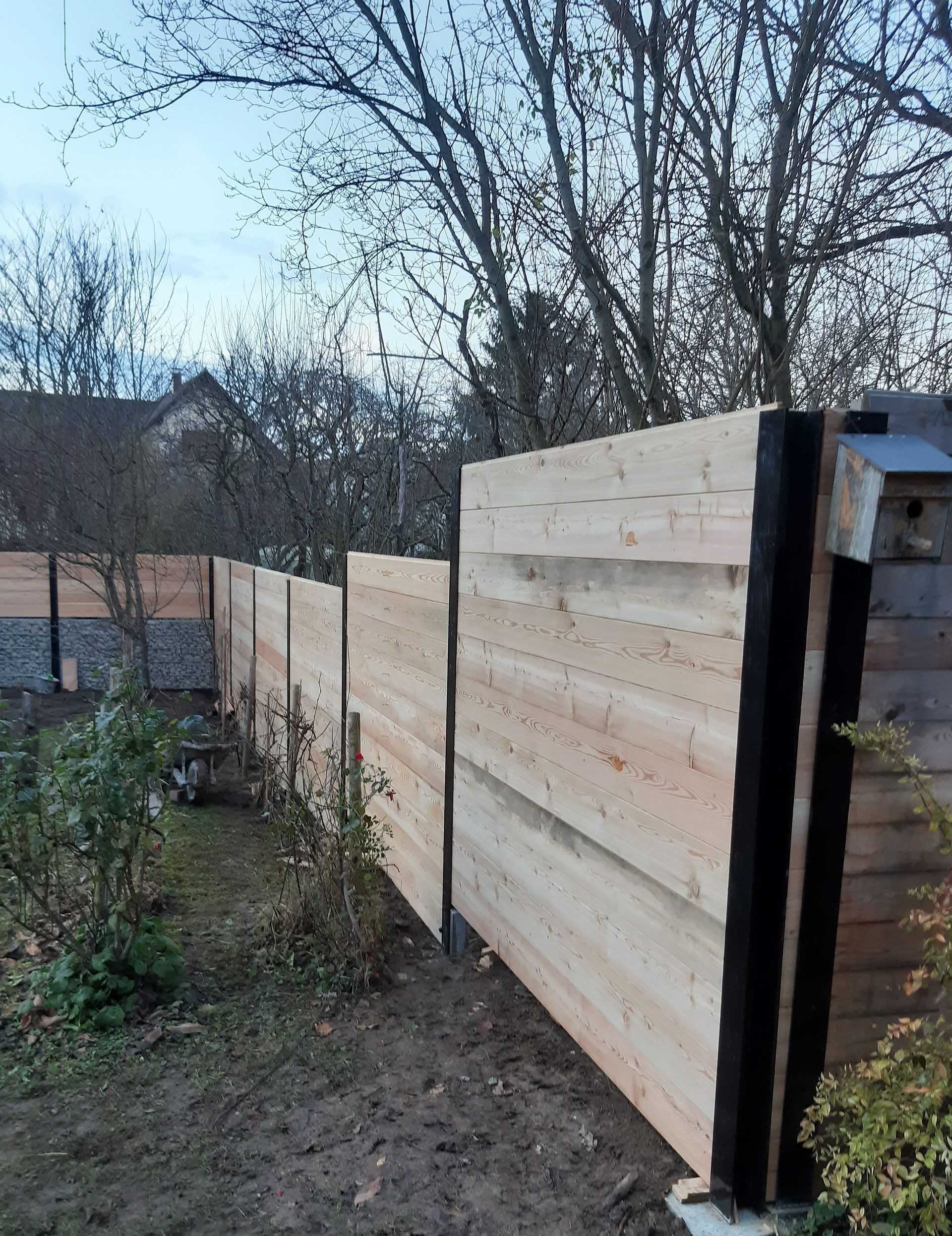 Clôture en bois dans une cour, avec en arrière-plan des arbres et une maison.