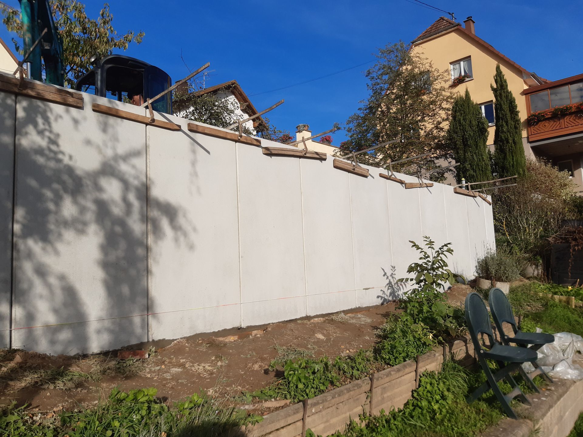 Construction d'un mur en béton gris dans un quartier résidentiel, avec des maisons et des arbres en arrière-plan.