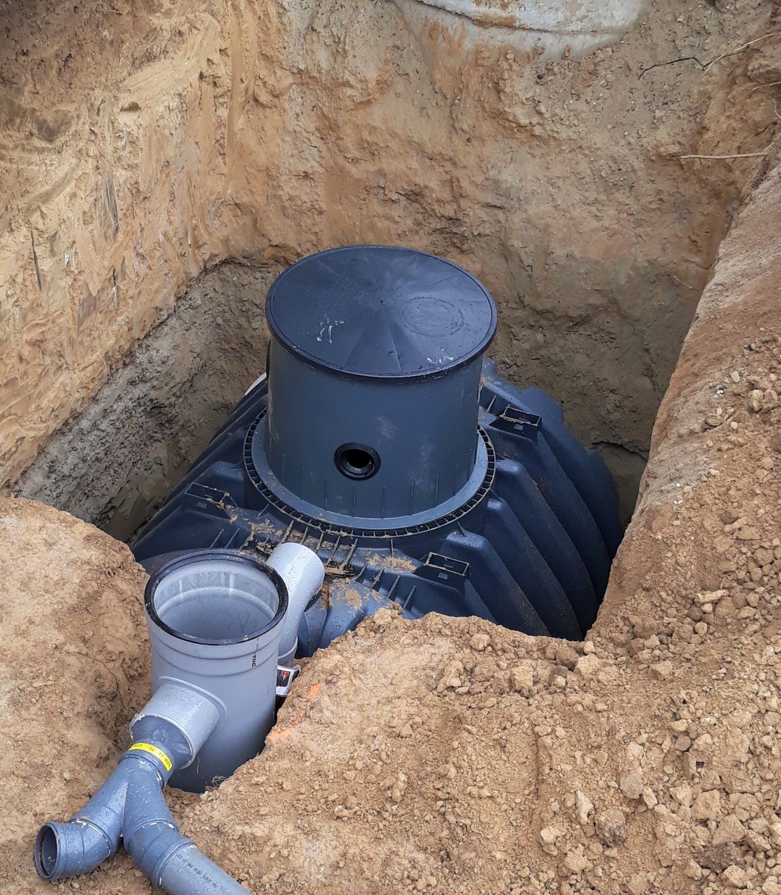 Installation d'une fosse septique dans une excavation carrée. Des tuyaux gris sont raccordés à une fosse noire nervurée.